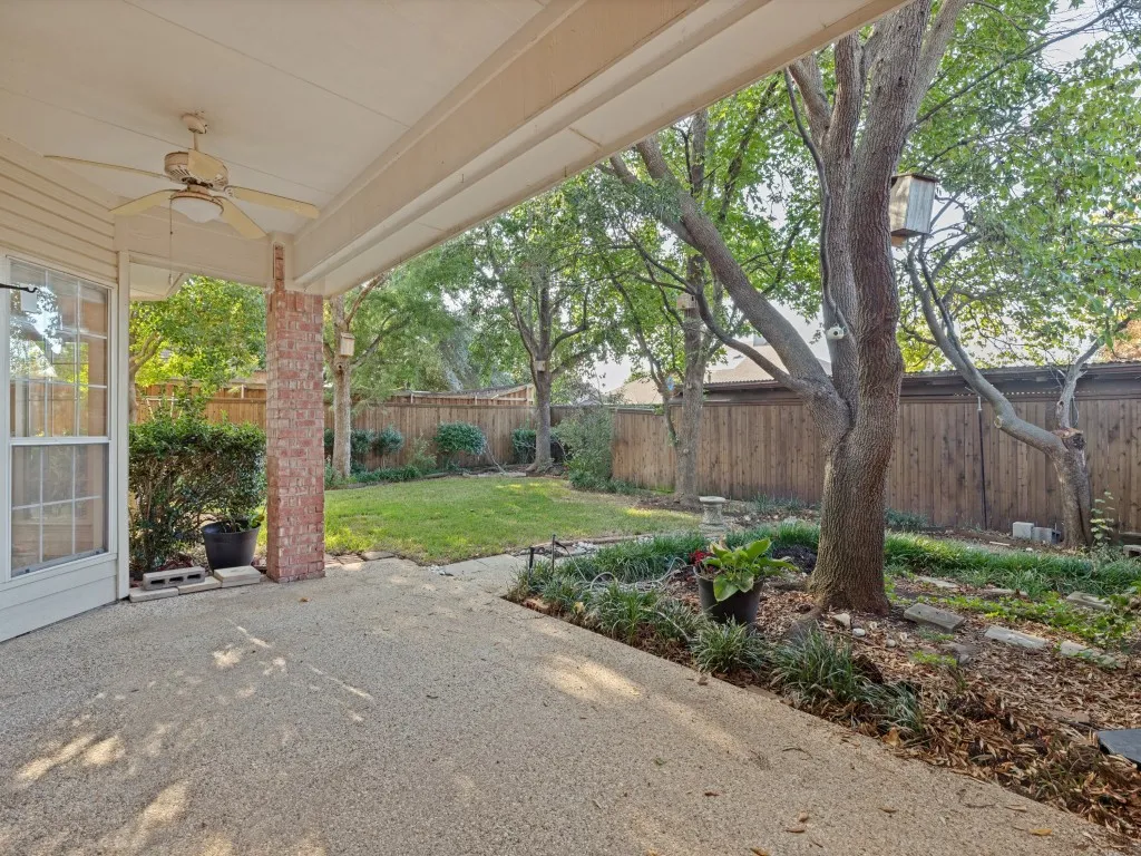 Covered patio featuring an exposed aggregate concrete surface, brick support column, and overhead ceiling fan, opening to a fenced backyard with established trees