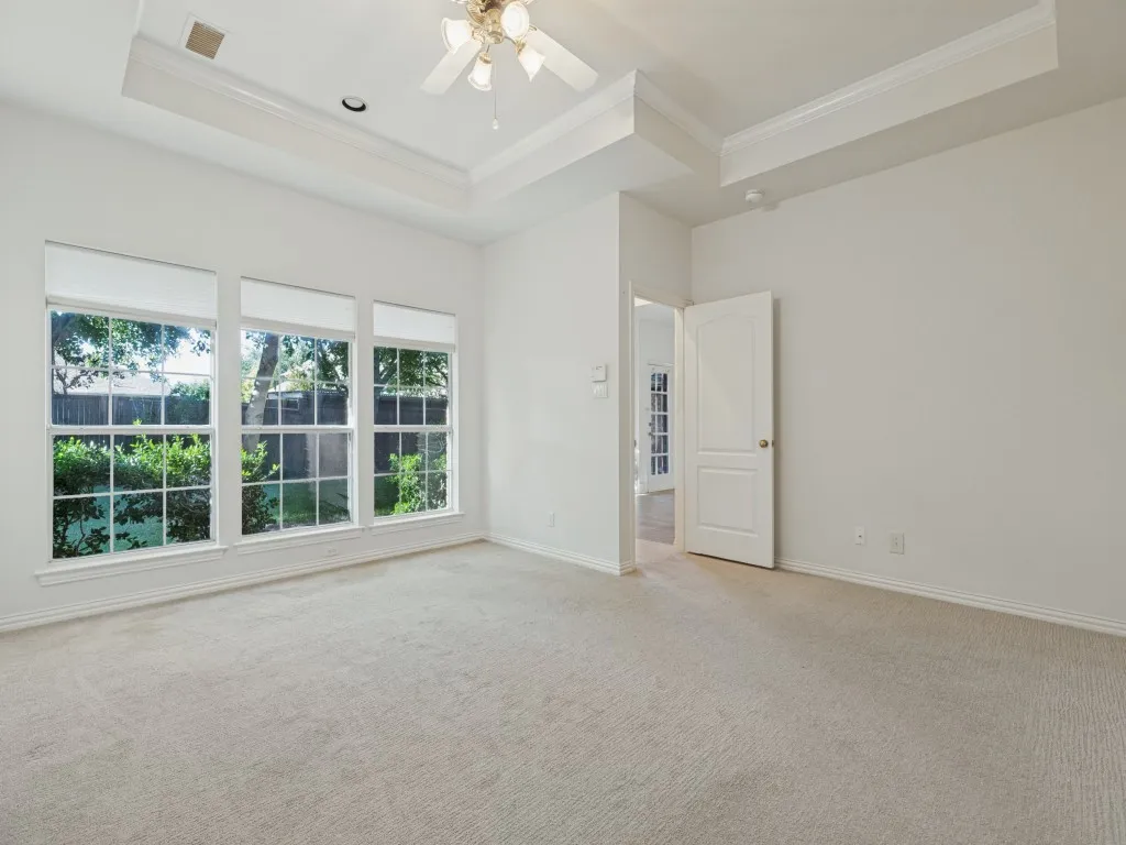 Tray ceiling with crown molding, three large windows with grid detailing, neutral carpeting, and a ceiling fan with integrated lighting