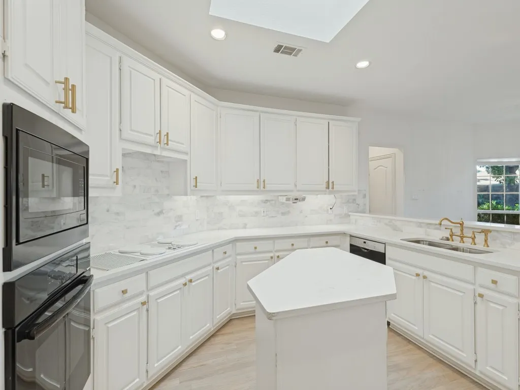 Kitchen featuring white cabinetry with gold-tone hardware, stone-surface countertops, stone-tile backsplash, built-in dual wall ovens, and a center island under a skylight