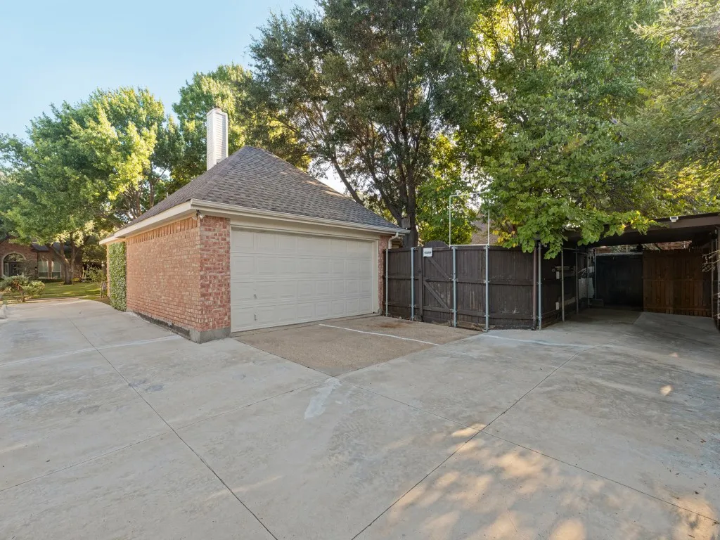 Detached brick garage featuring a multi-pitched shingled roof, chimney, concrete driveway, and attached metal-framed carport