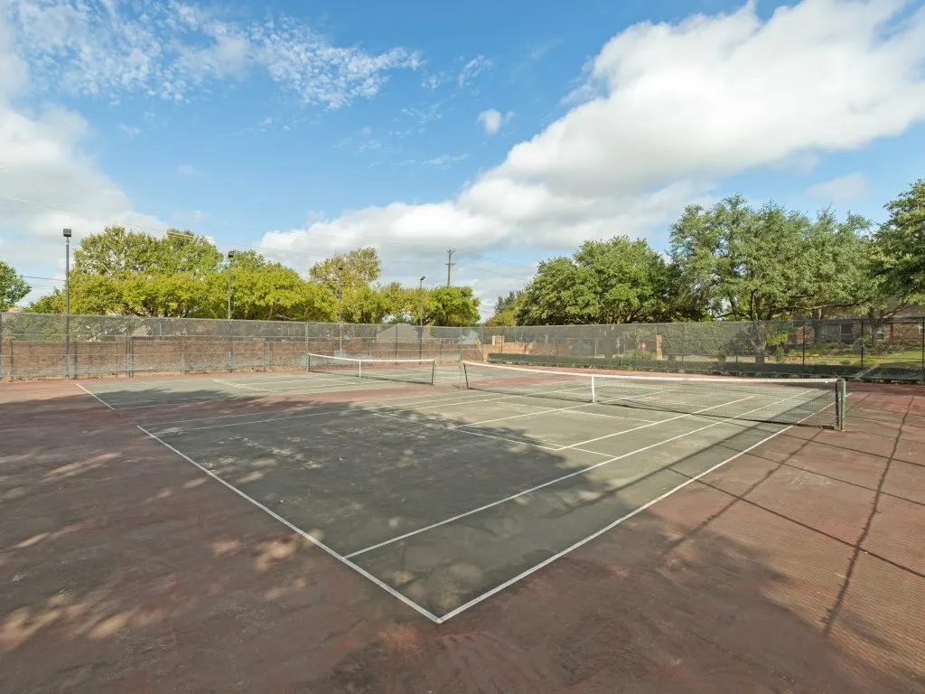 Dual outdoor tennis courts featuring perimeter chain-link fencing, structural lighting poles, and mature tree boundaries