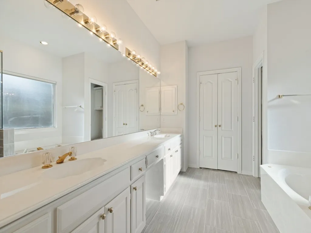 Bathroom featuring a dual-sink vanity with a countertop, expansive wall-to-wall mirror, multi-bulb overhead lighting, and porcelain tile flooring