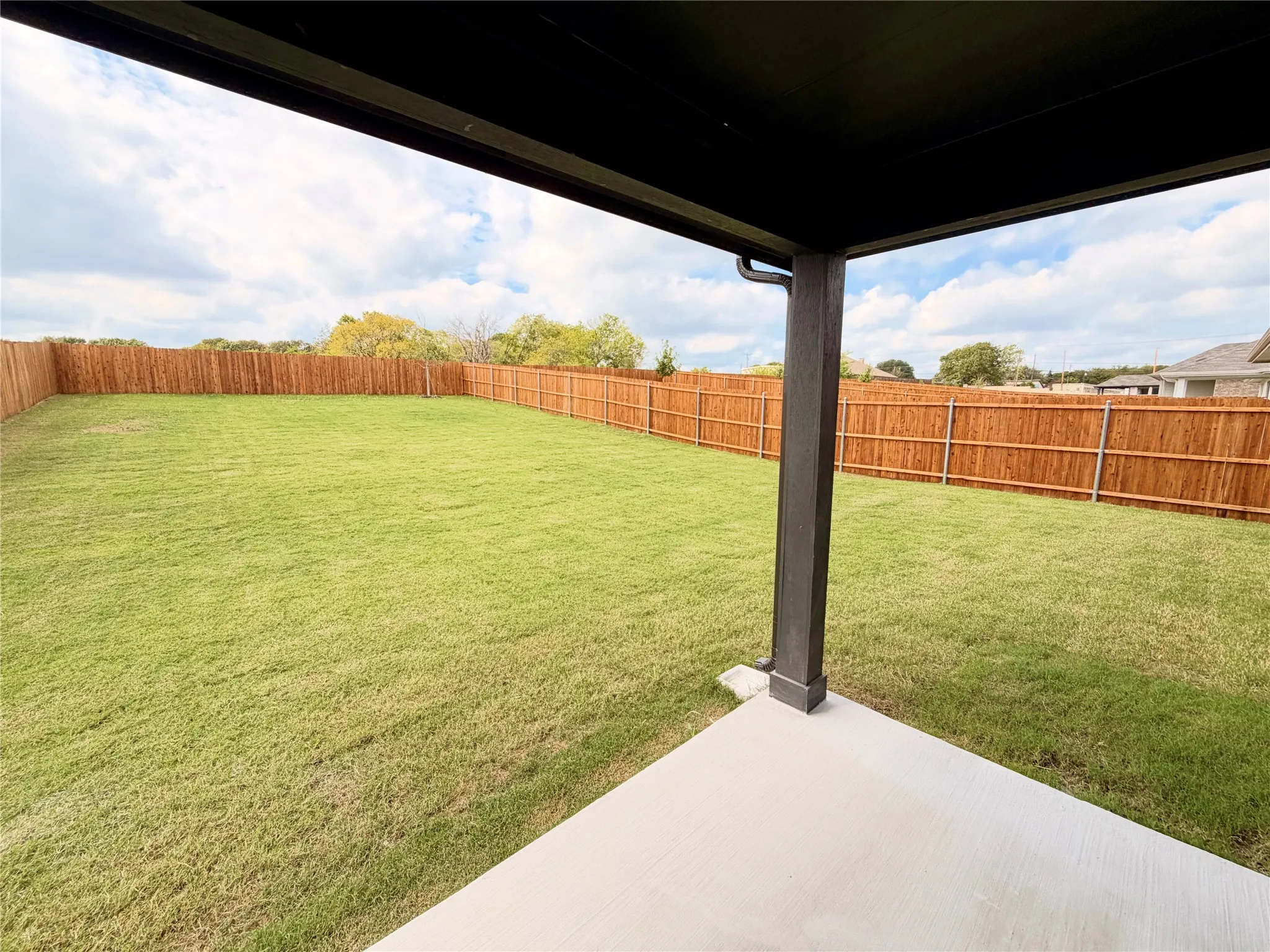 Covered concrete patio overlooking an expansive grass lawn enclosed by a tall wood privacy fence