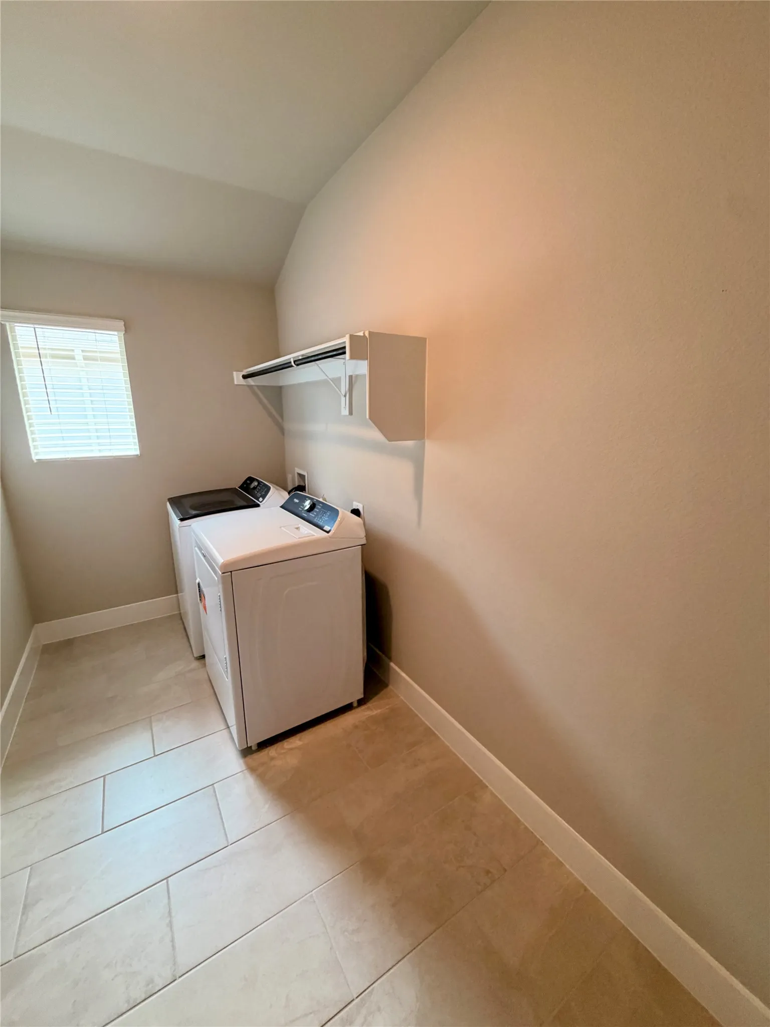 Laundry area featuring large-format ceramic tile flooring, a wall-mounted white storage shelf with integrated rod, and a window with horizontal blinds