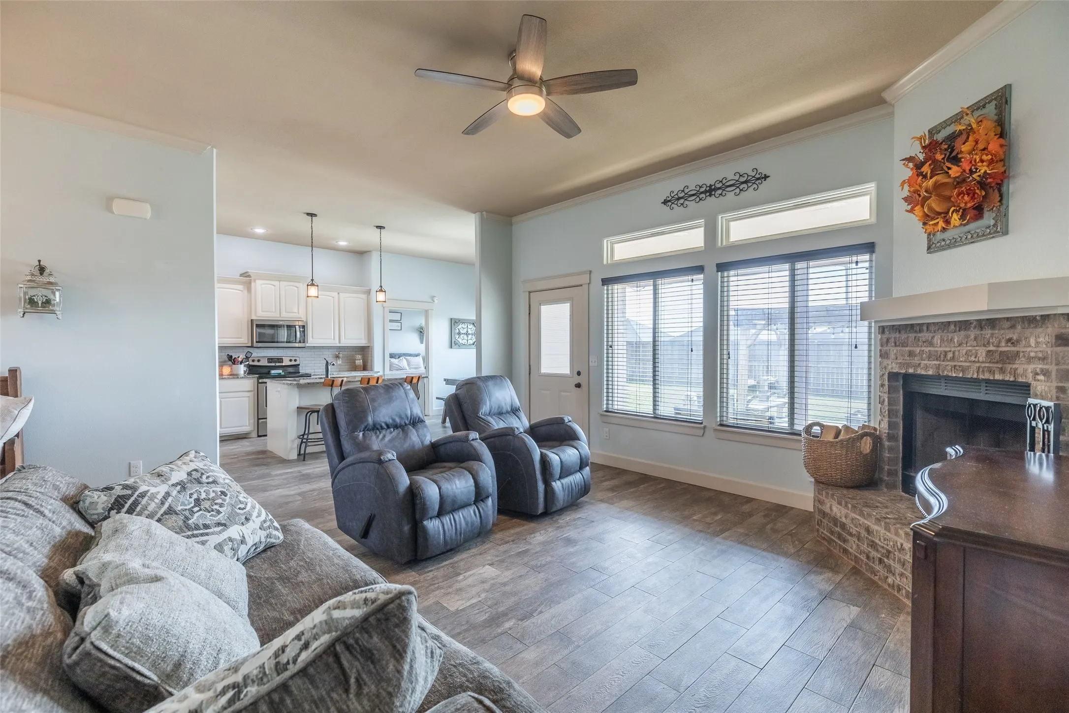 Open-concept interior featuring wood-finish flooring, a brick masonry fireplace, large windows with transoms, and a kitchen equipped with cabinetry and pendant lighting