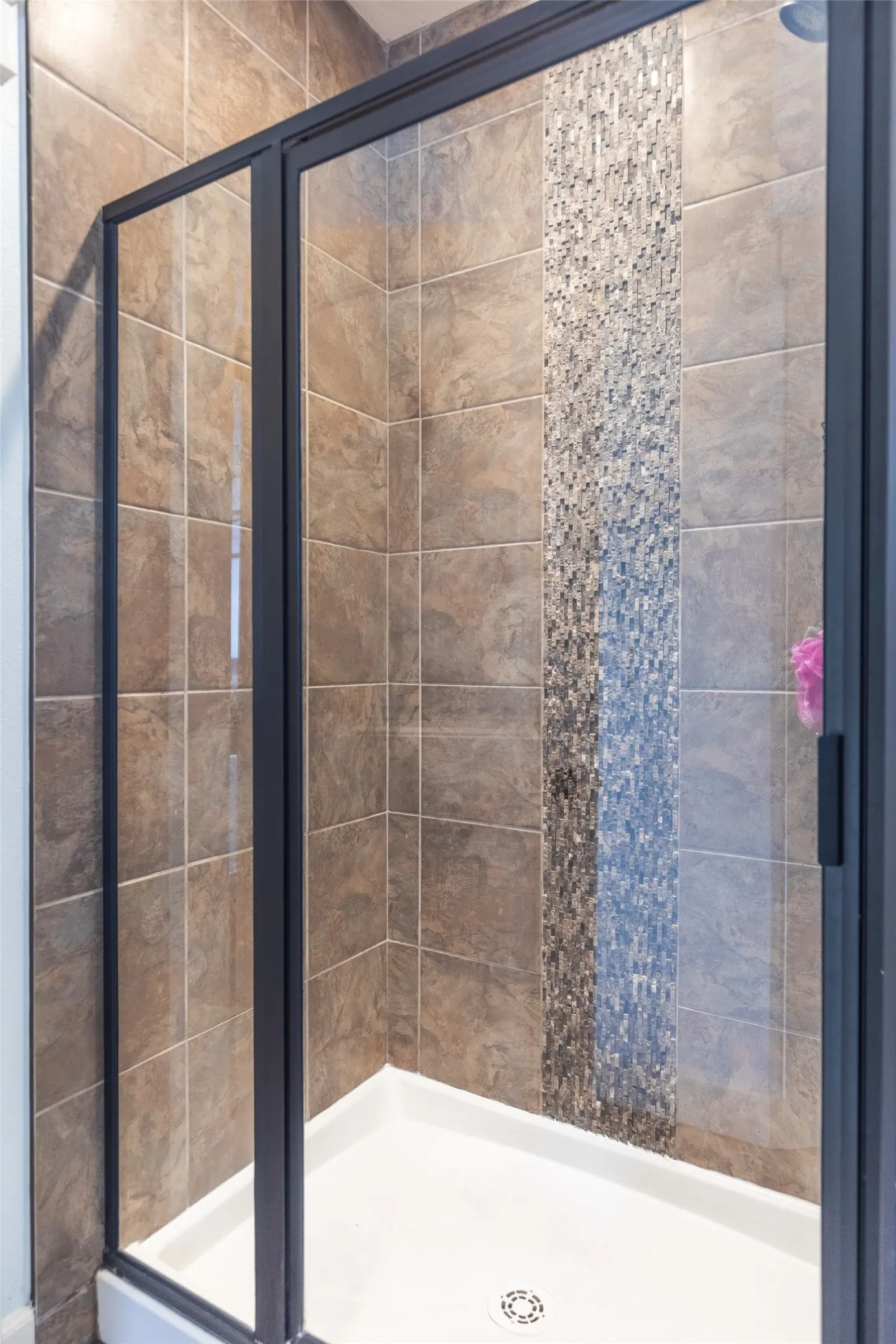 Shower enclosure featuring large-format ceramic wall tile, vertical glass mosaic tile inlay, white acrylic shower pan, and black metal-framed glass doors