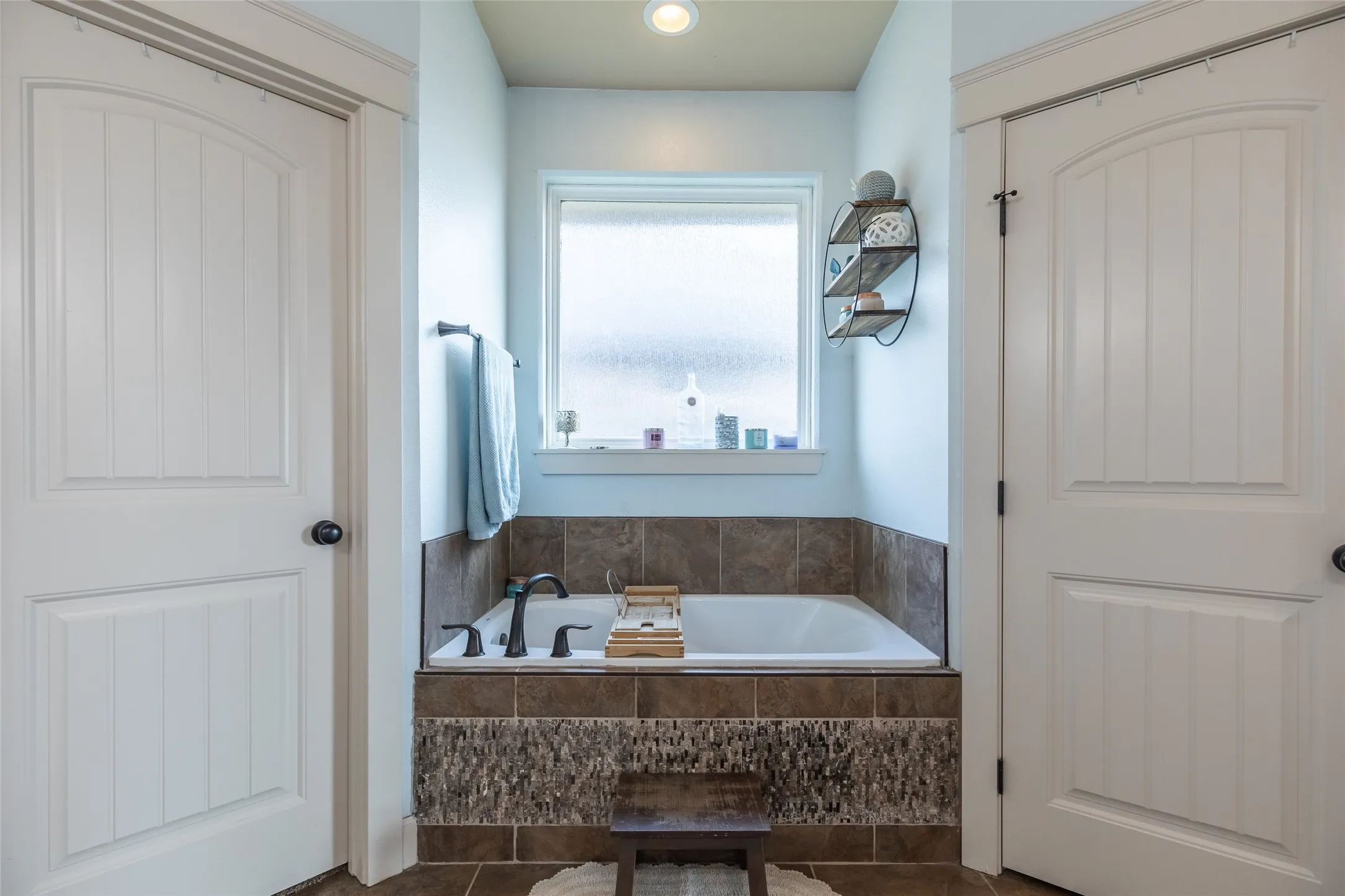 Inset soaking tub featuring a stone-tiled surround, dark metallic faucet fixtures, privacy-glass window, and two-panel interior doors with casing