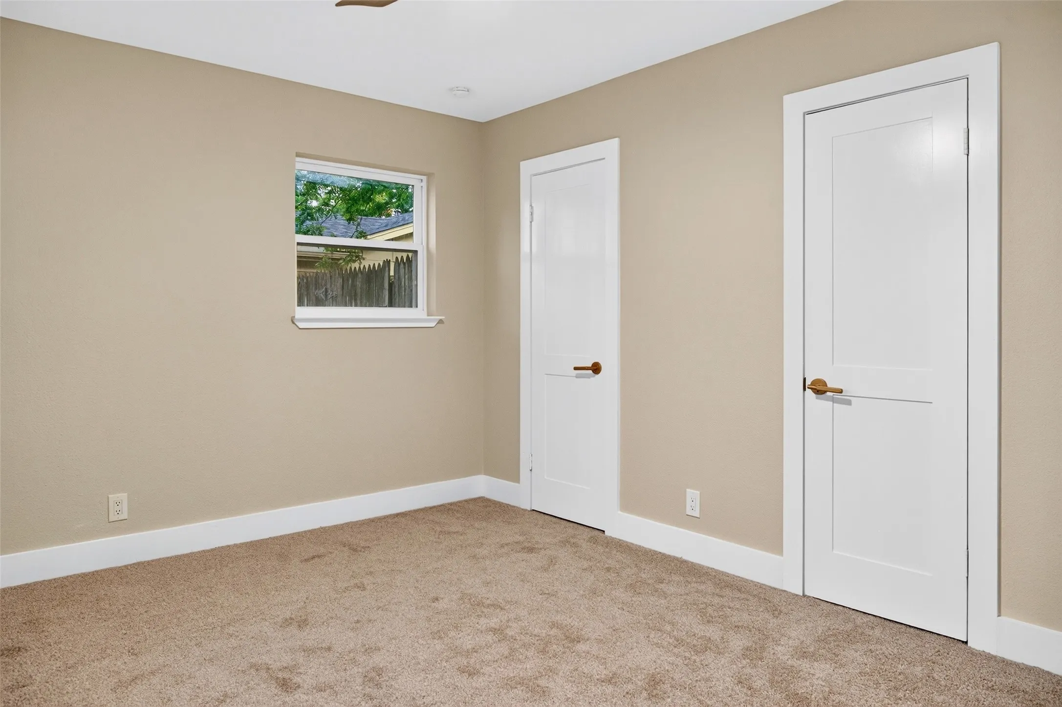 Neutral-toned interior room featuring wall-to-wall carpeting, two white shaker-style doors with metallic hardware, and a single-pane window