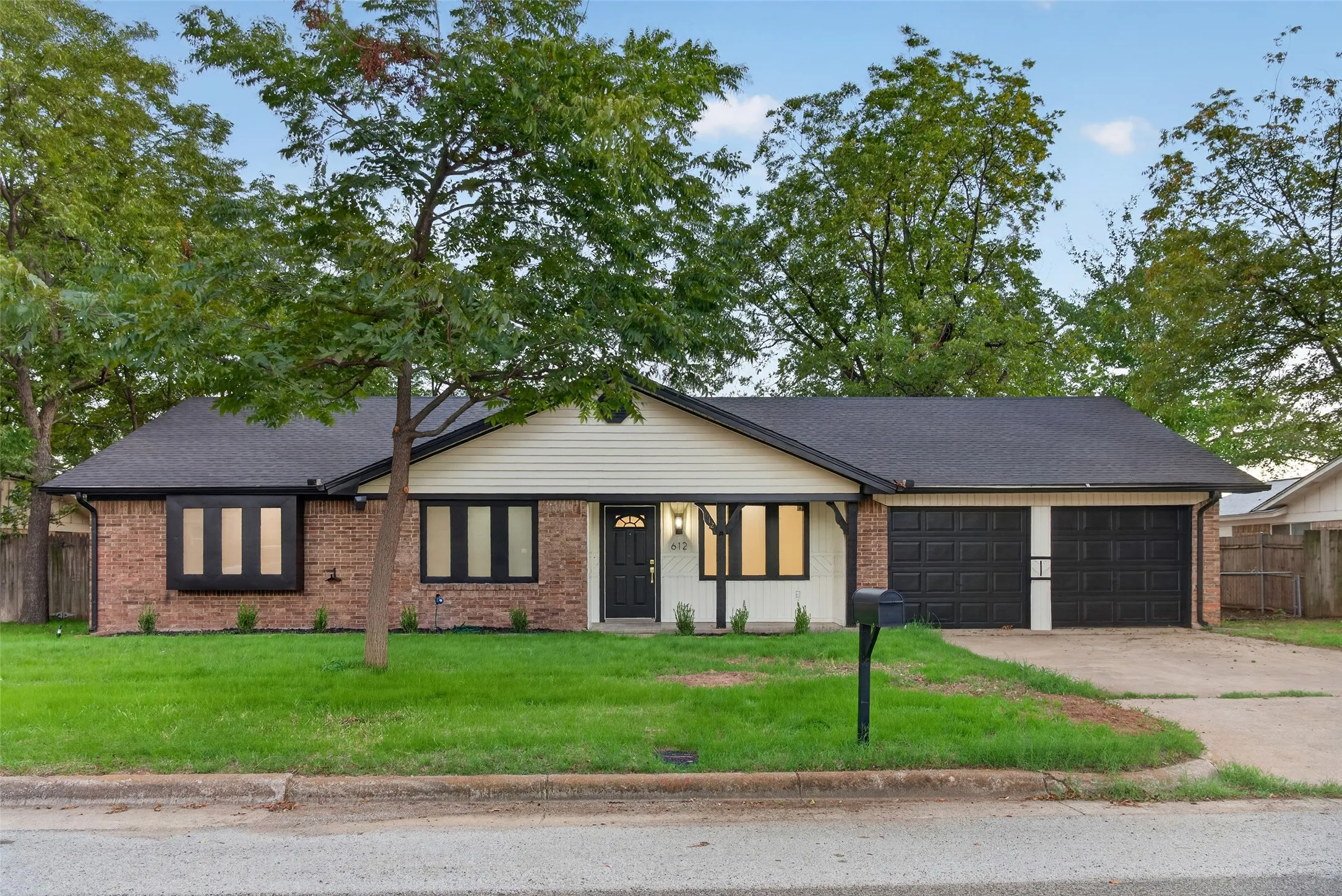 Single-story exterior featuring a brick and siding facade, a covered entryway, black-trimmed windows, a two-car garage, and mature trees