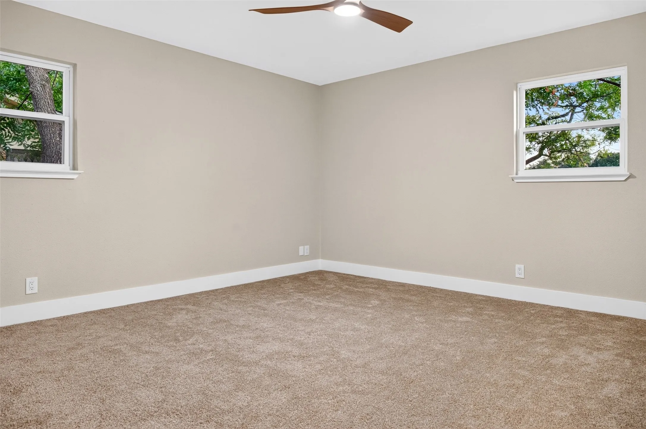 Neutral-toned interior featuring wall-to-wall carpeting, dual operable windows with exterior foliage, and a ceiling-mounted fan with integrated lighting