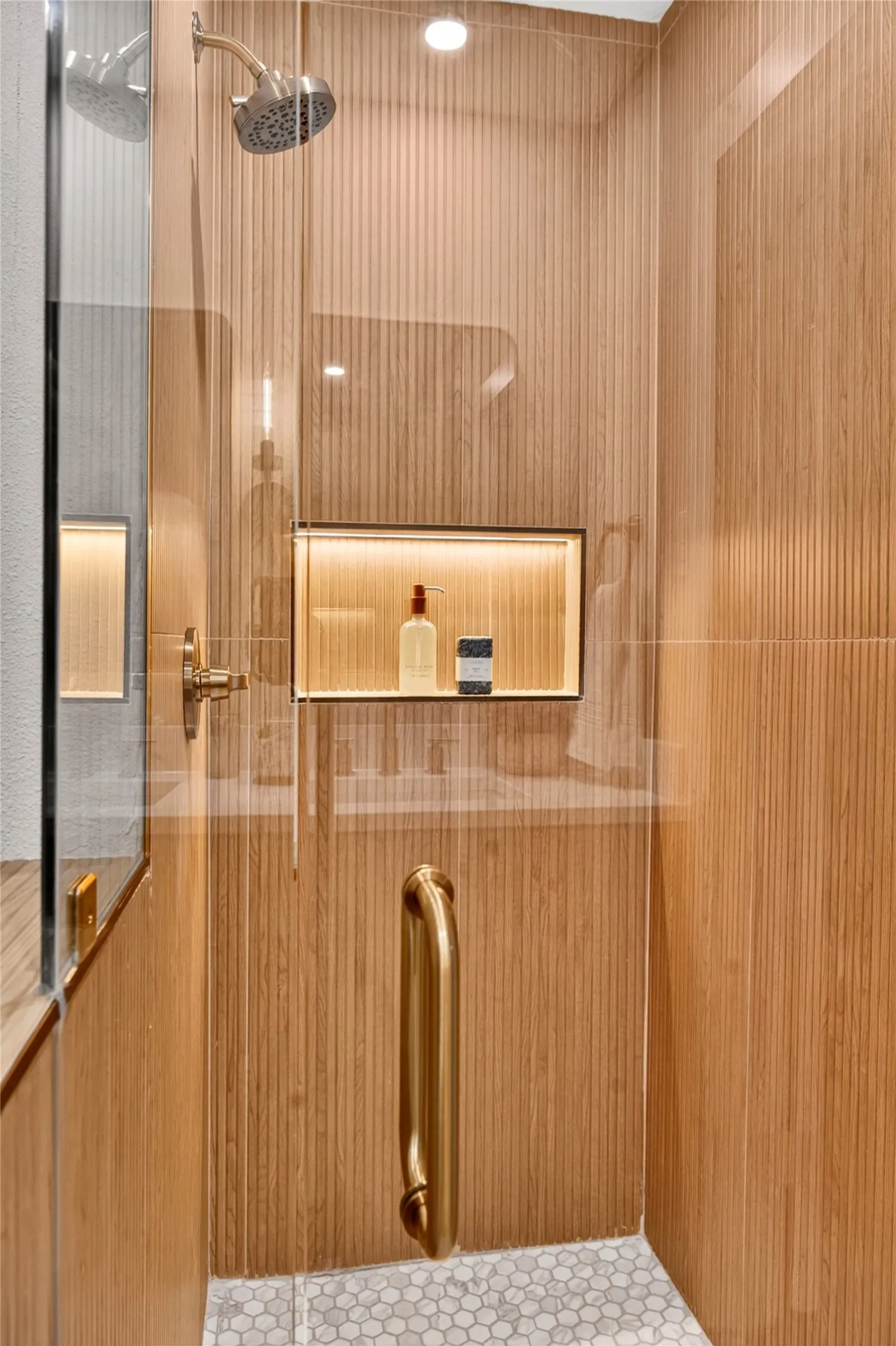 Walk-in shower featuring vertically ribbed wood-finish wall tile, frameless glass door, metallic shower fixture, integrated recessed shelving with accent lighting, and hexagonal tile flooring