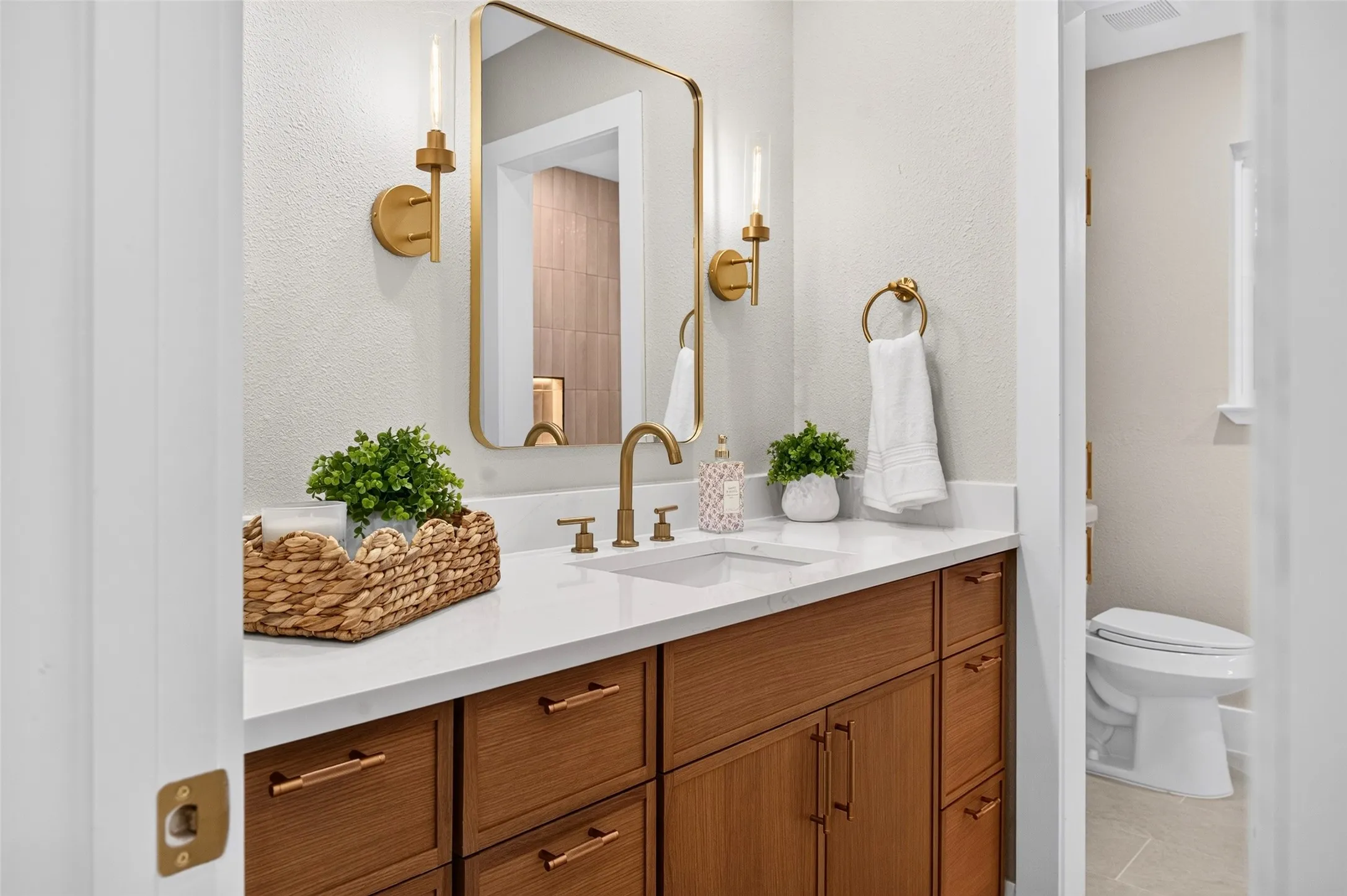 Wood-finish vanity cabinetry with brushed gold hardware, quartz countertop, rectangular undermount sink, matching gold-tone wall-mounted light fixtures, and a framed gold-finished mirror