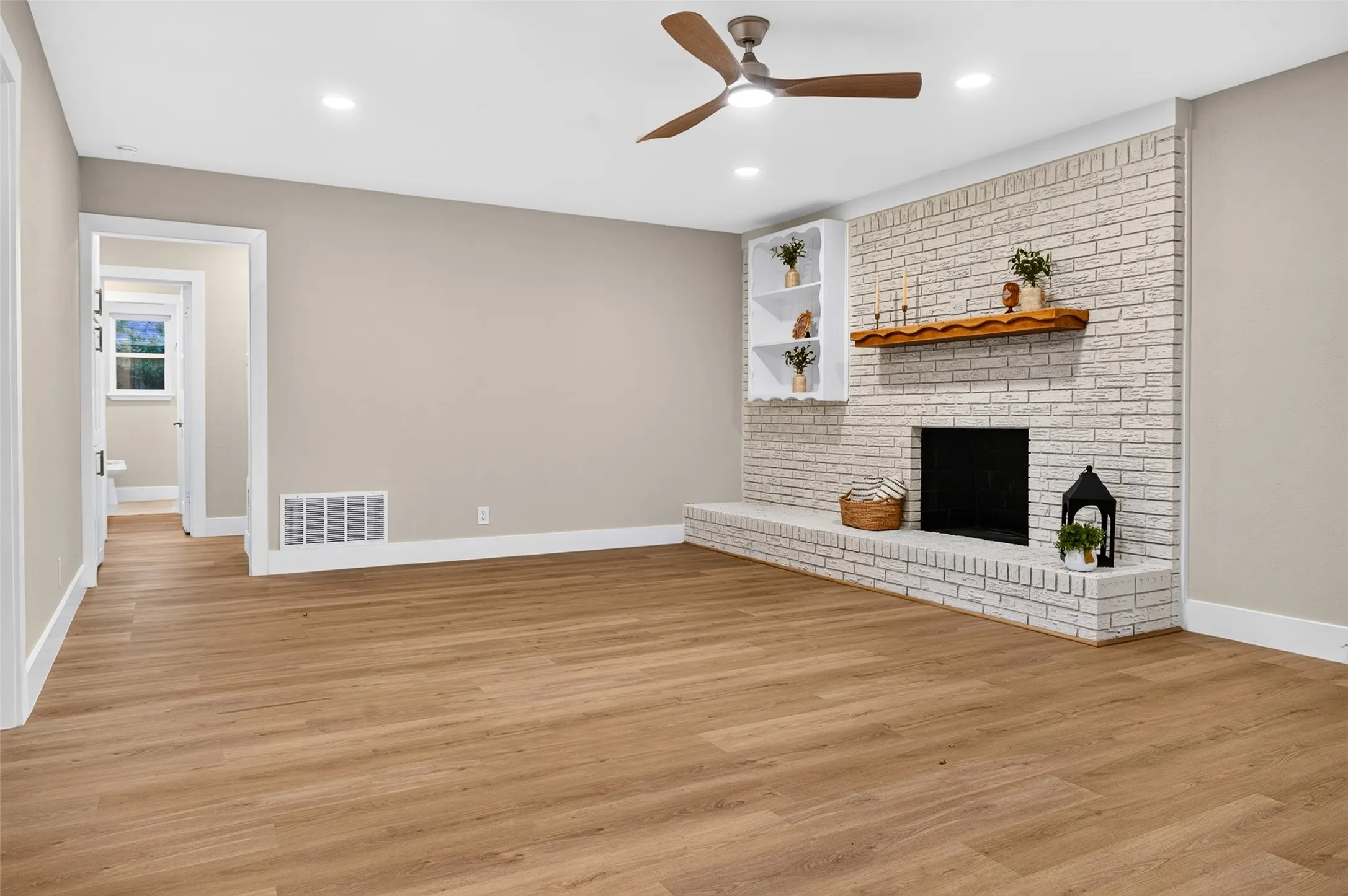 Painted brick fireplace with a wood mantel, wood-finish flooring, built-in shelving, and a modern ceiling fan