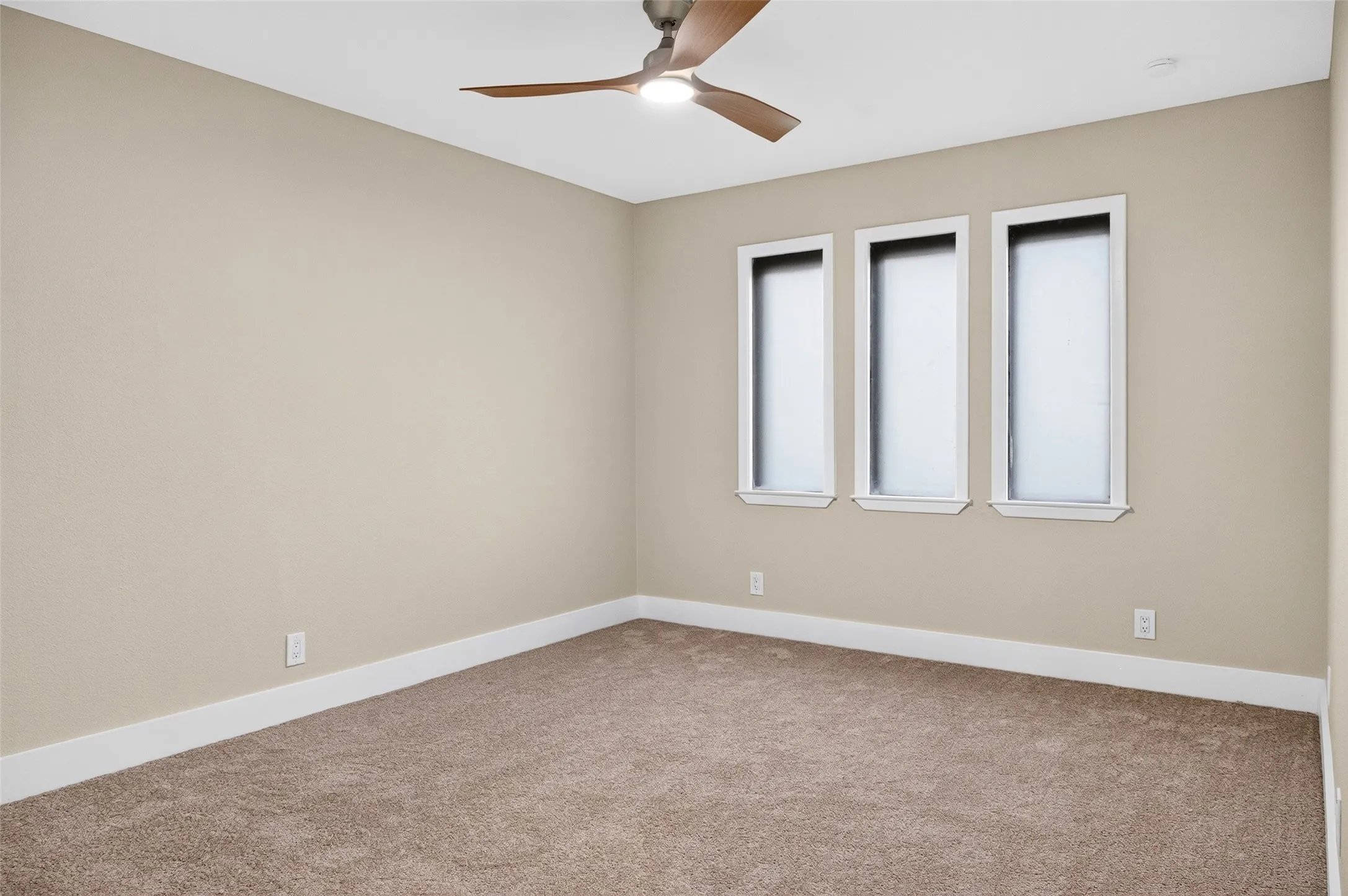 Neutral-toned interior room featuring wall-to-wall carpeting, three vertical windows, and a ceiling-mounted light fixture with wood-finish blades