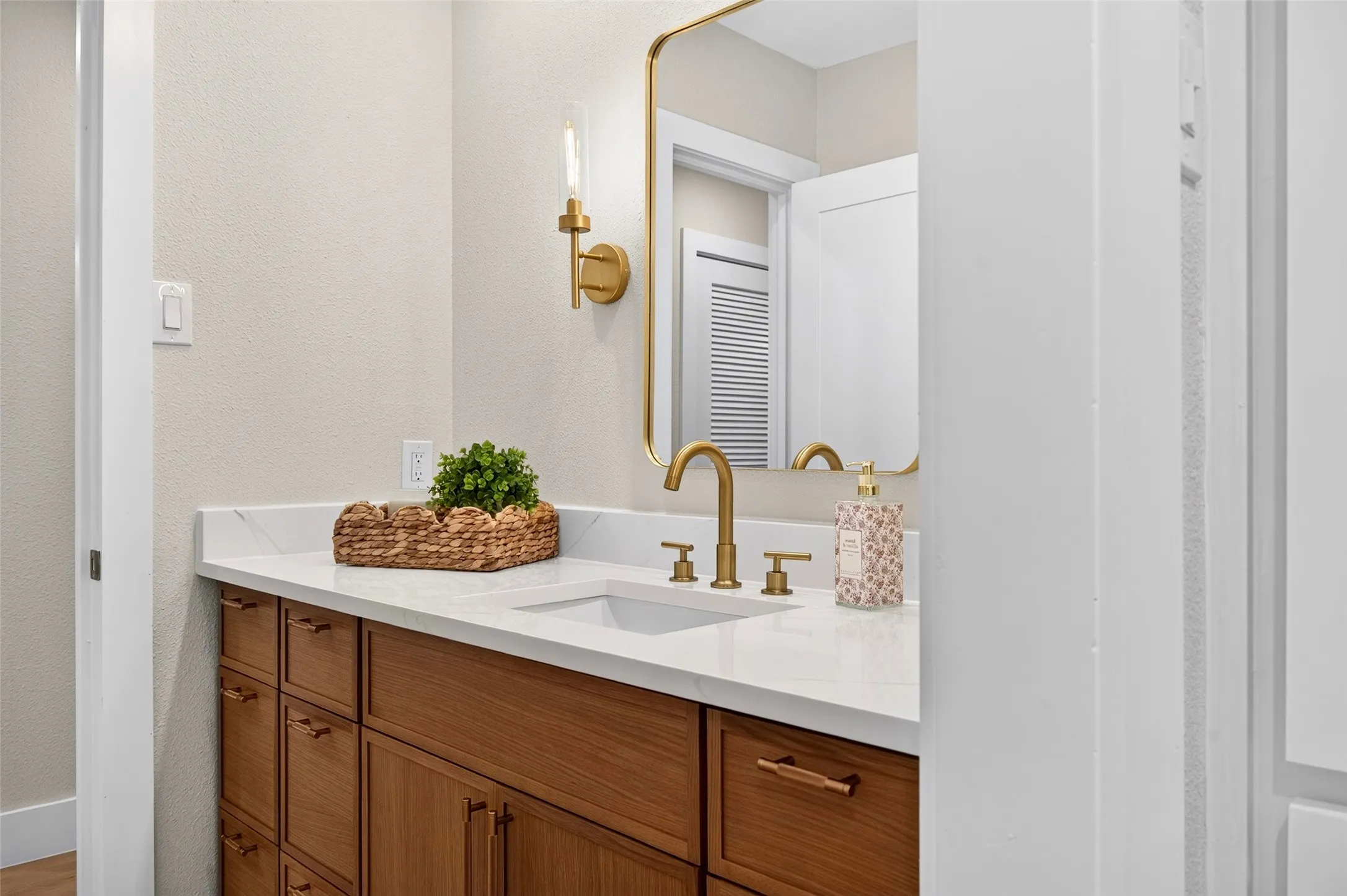 Wood-finish cabinetry, quartz countertop, rectangular undermount sink, brass-finish widespread faucet, and matching brass-rimmed mirror