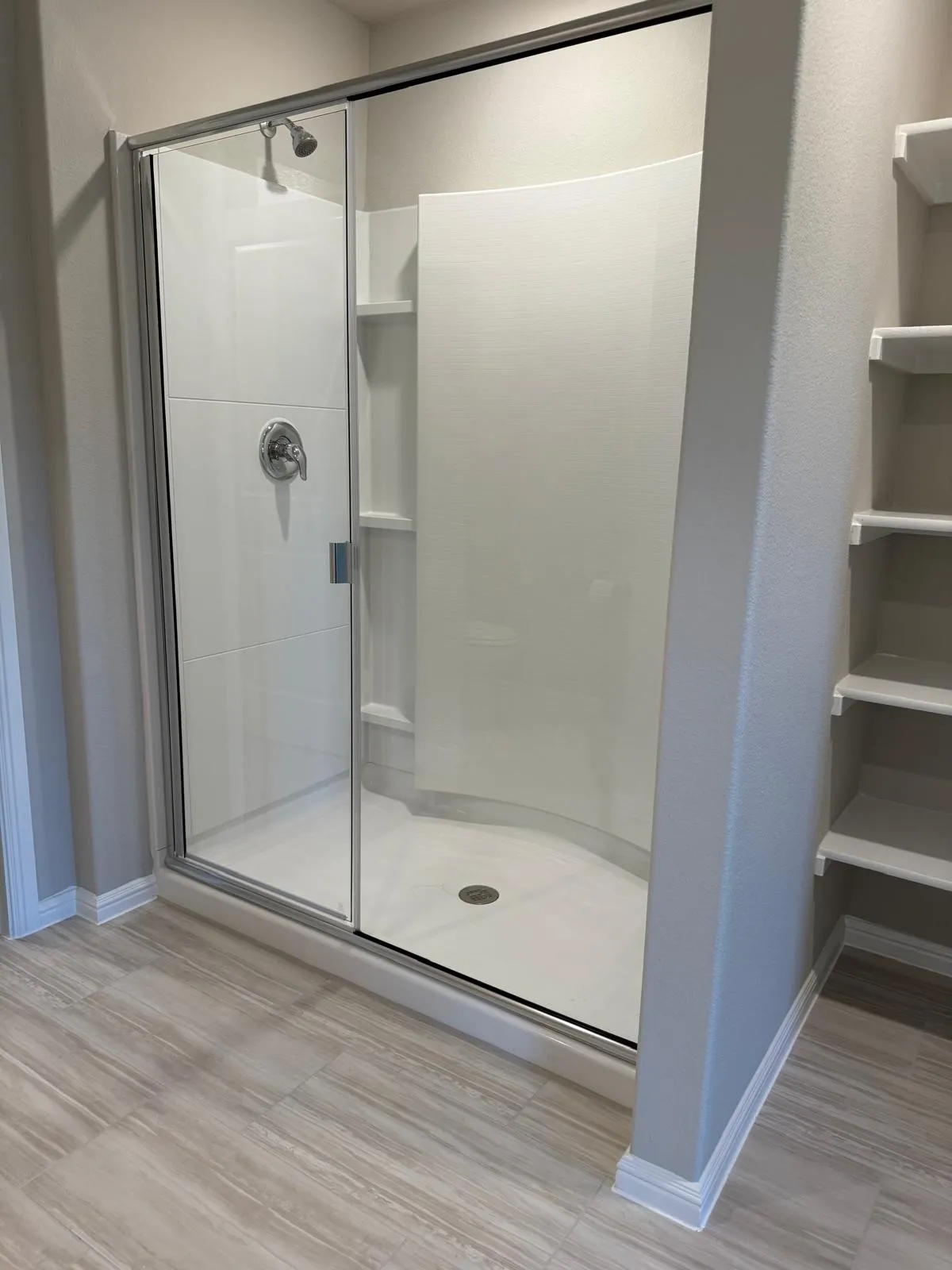 Shower enclosure with a curved glass sliding door, integrated shelving, a single-handle faucet fixture, and wood-finish tile flooring, accompanied by built-in wall shelving