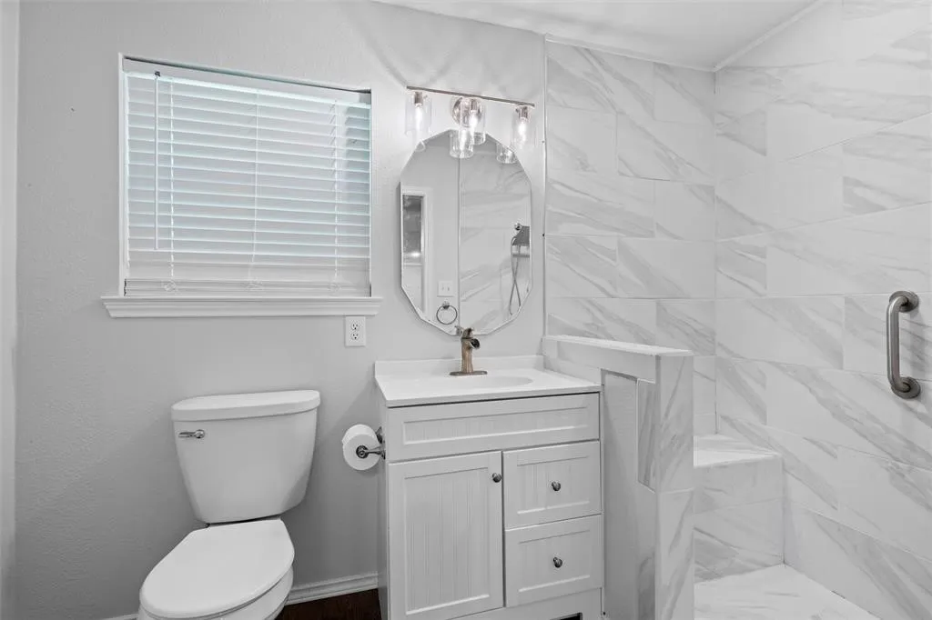 Bathroom featuring a white vanity cabinet with an integrated countertop, oval wall-mounted mirror, three-light fixture, and a tiled shower stall with a built-in bench