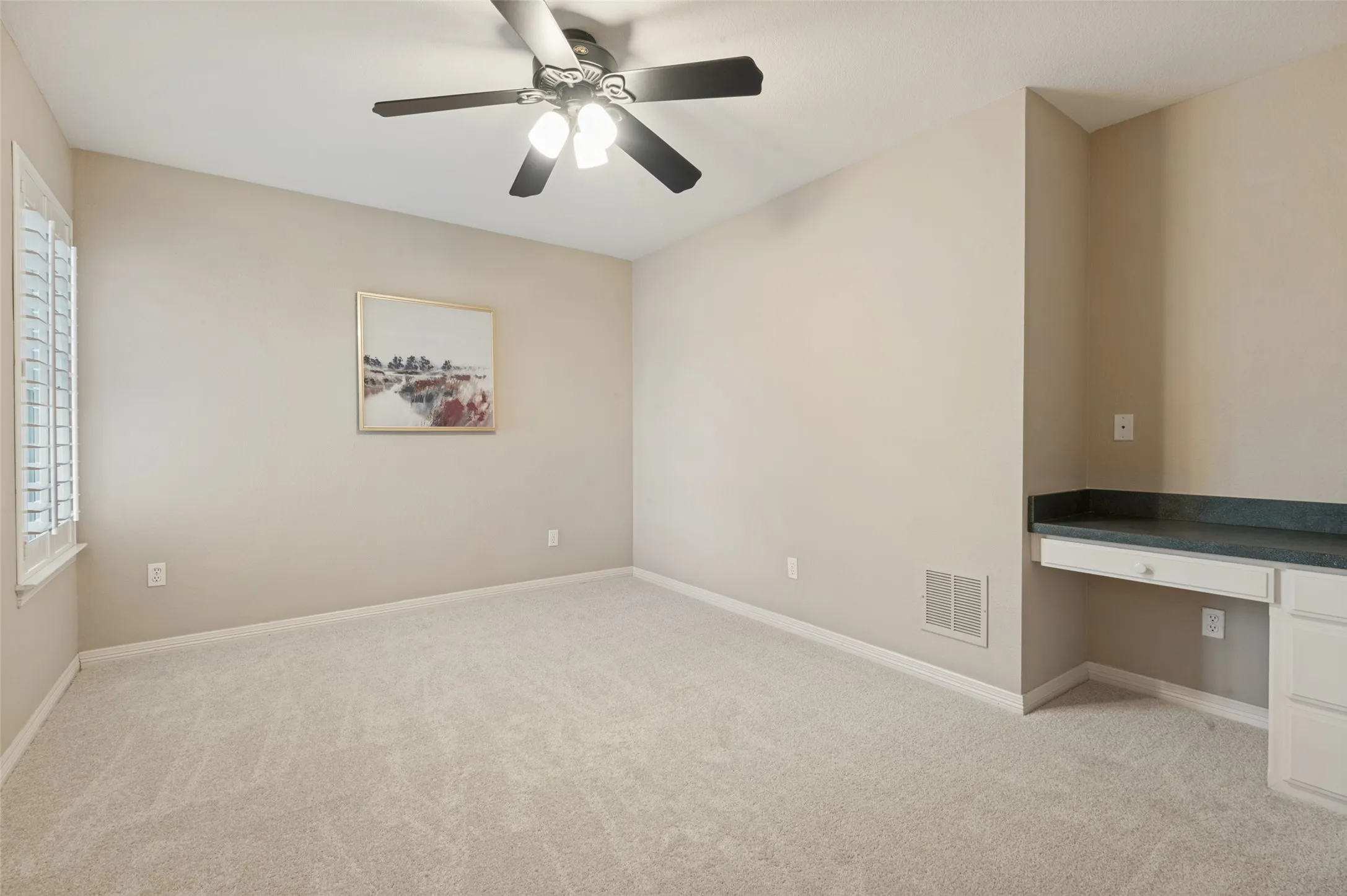 Carpeted interior space featuring a ceiling fan with integrated lighting, plantation shutters, and a built-in desk with stone-finish countertop and cabinetry