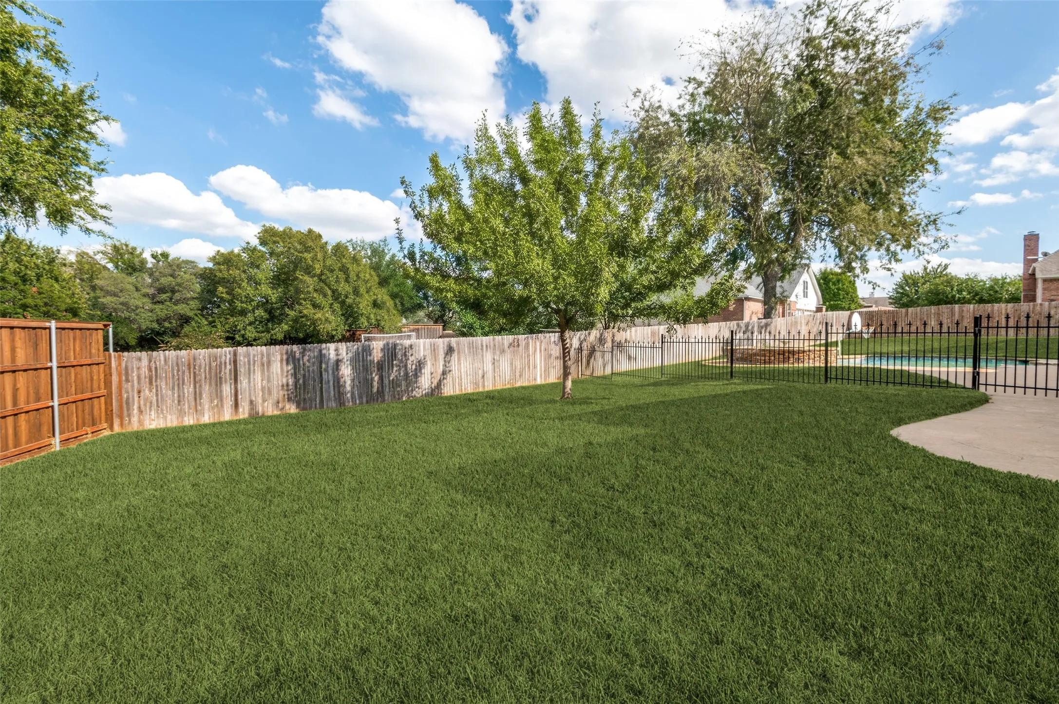 Backyard featuring a manicured lawn, wood privacy fencing, a metal perimeter fence, concrete hardscaping, and mature trees