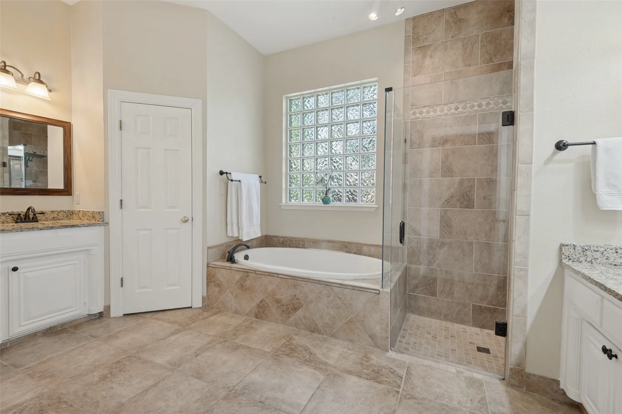 Dual vanity area featuring stone-look countertops, soaking tub with tile surround, glass-enclosed walk-in shower, and glass block window