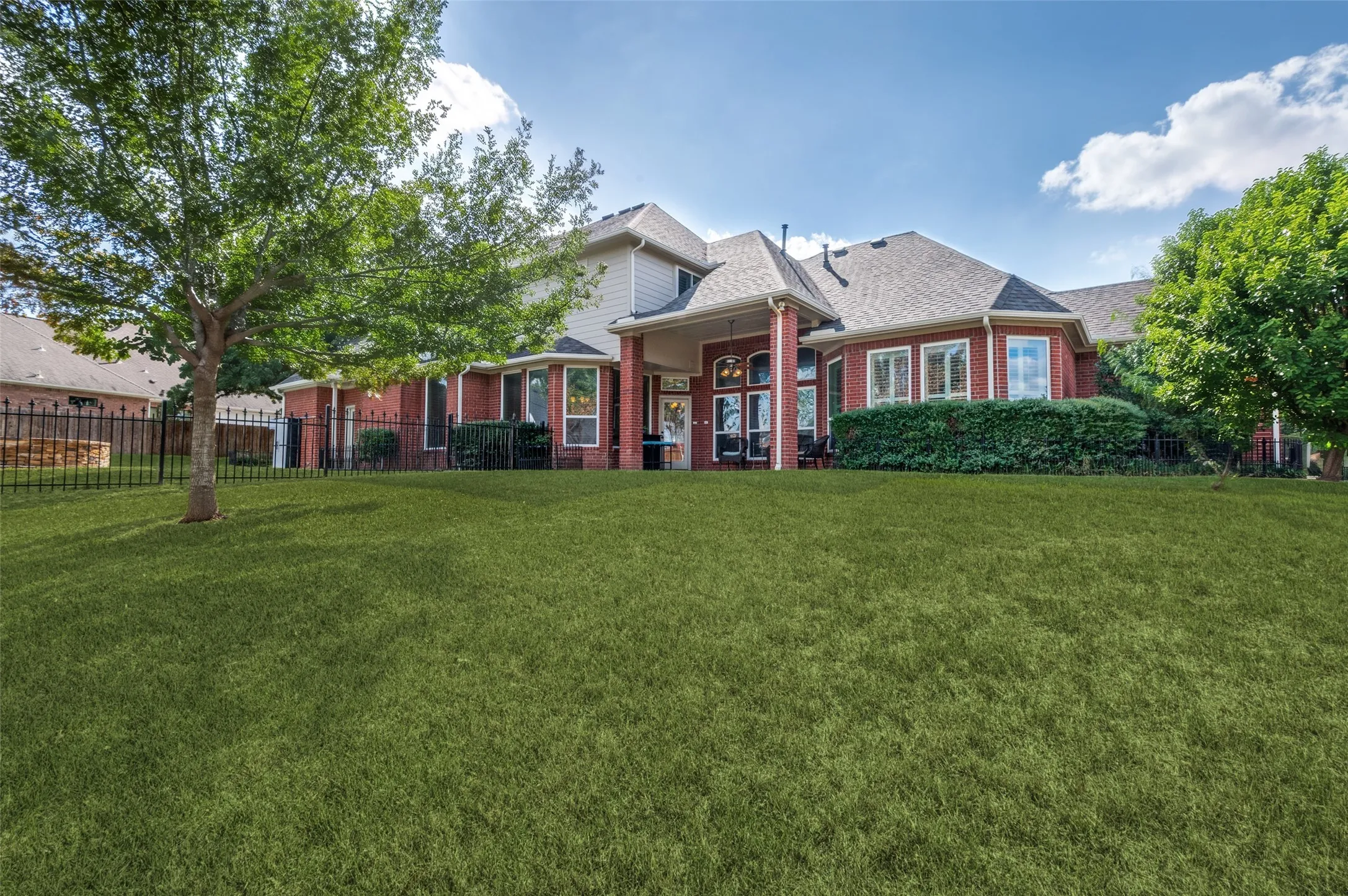Expansive backyard featuring a manicured lawn, mature trees, metal perimeter fencing, brick-and-siding exterior, and a covered patio
