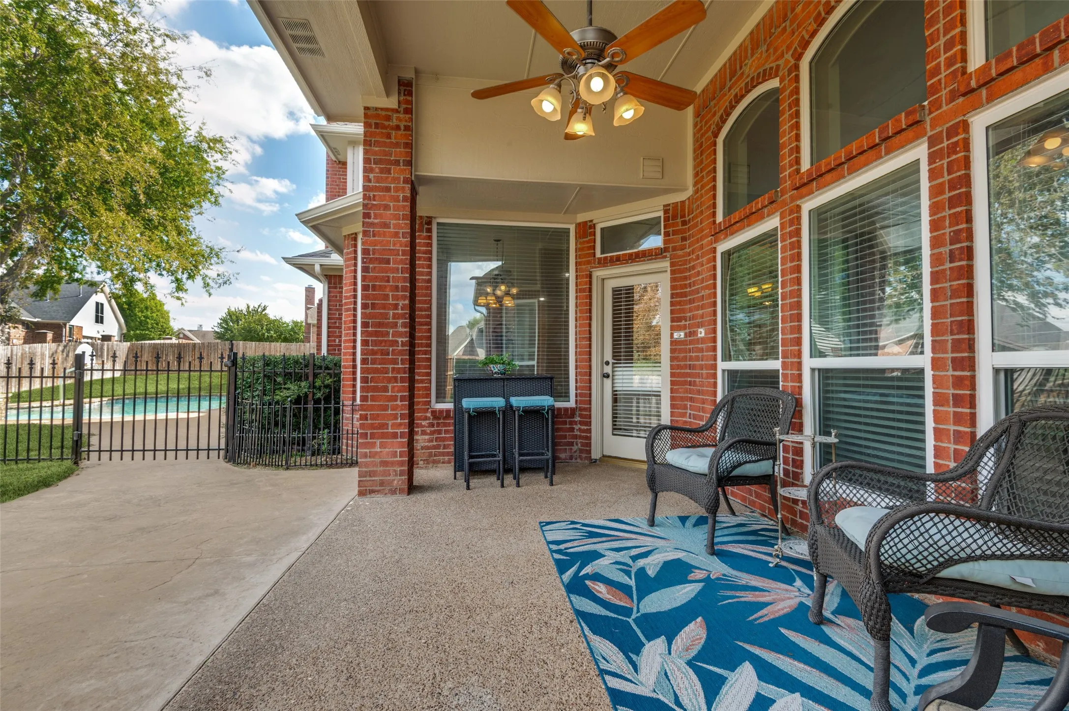 Covered patio featuring brick exterior walls, aggregate concrete flooring, an overhead ceiling fan with integrated lighting, and metal perimeter fencing
