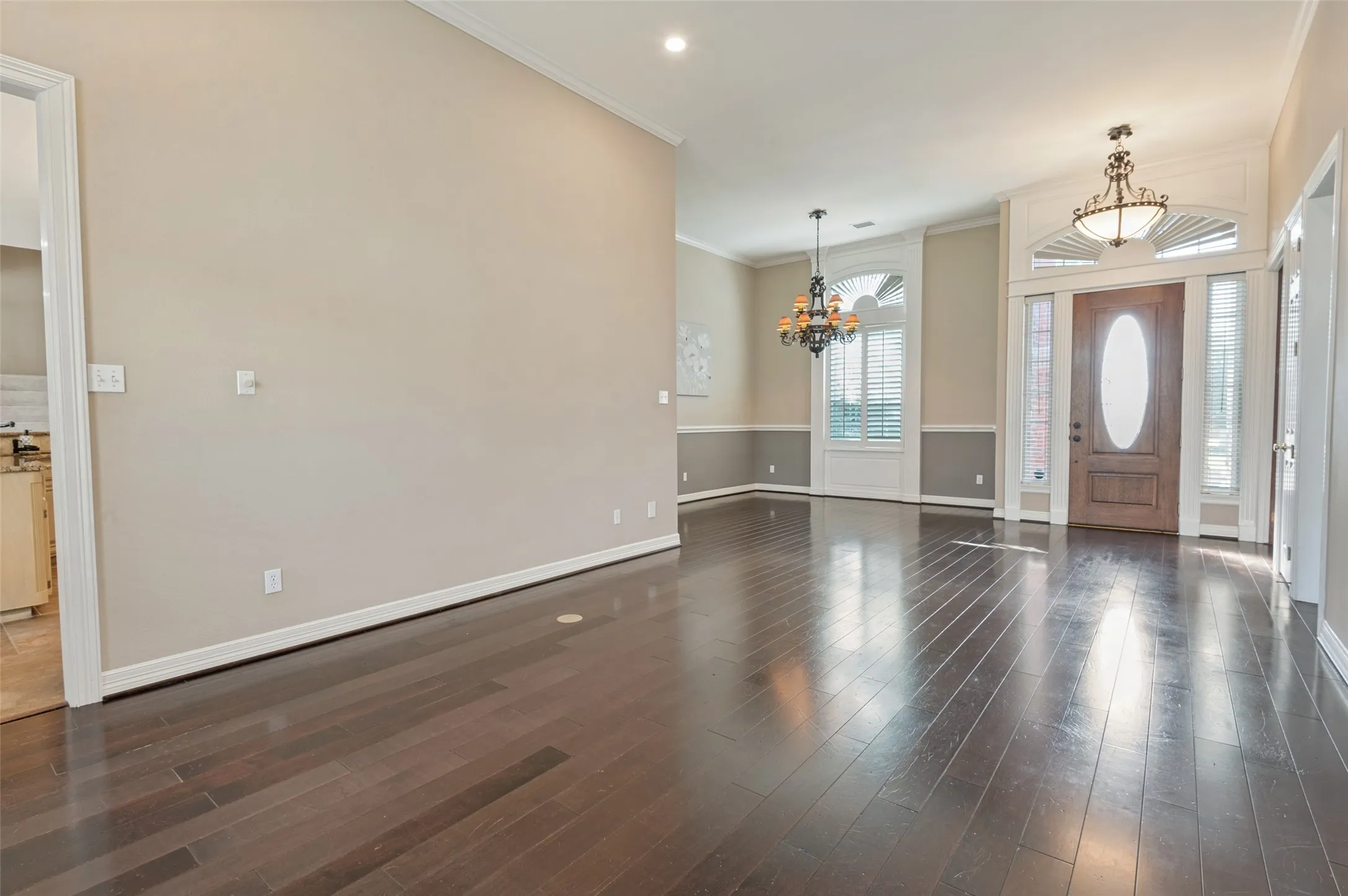 Dark wood-finish flooring, high ceilings with crown molding, decorative chandeliers, arched windows with plantation shutters, and a wood entry door with an oval glass insert and transom window