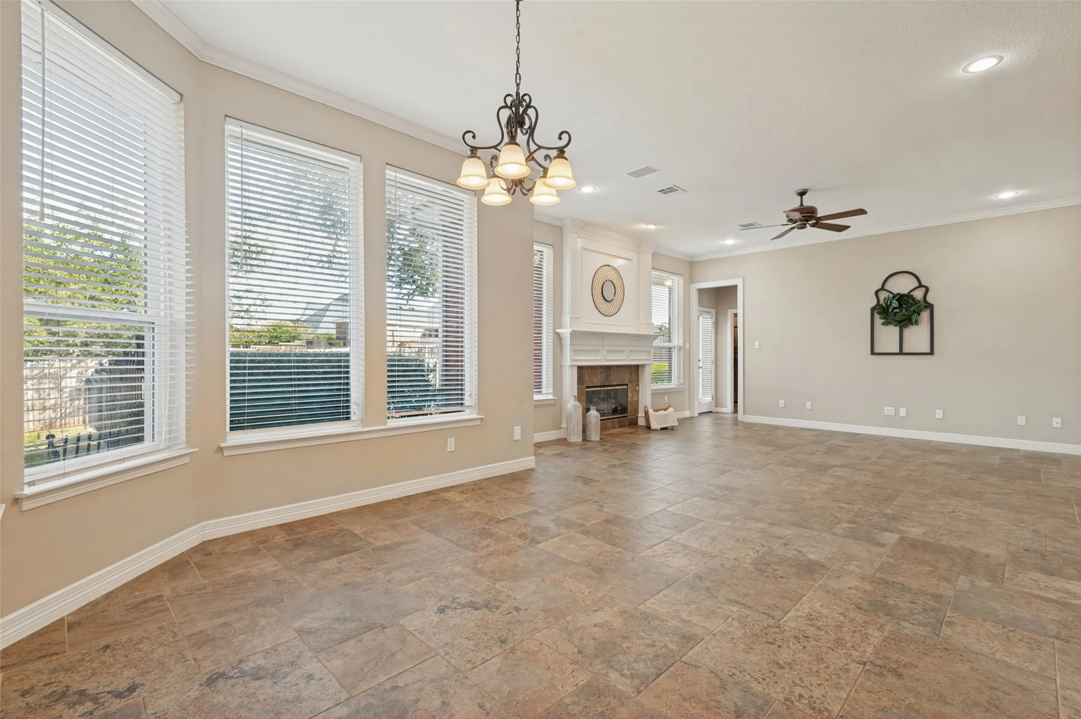 Open-concept room featuring large-format tile flooring, a stone-surround fireplace with a classic mantel, oversized windows with blinds, a multi-light chandelier, and a ceiling fan