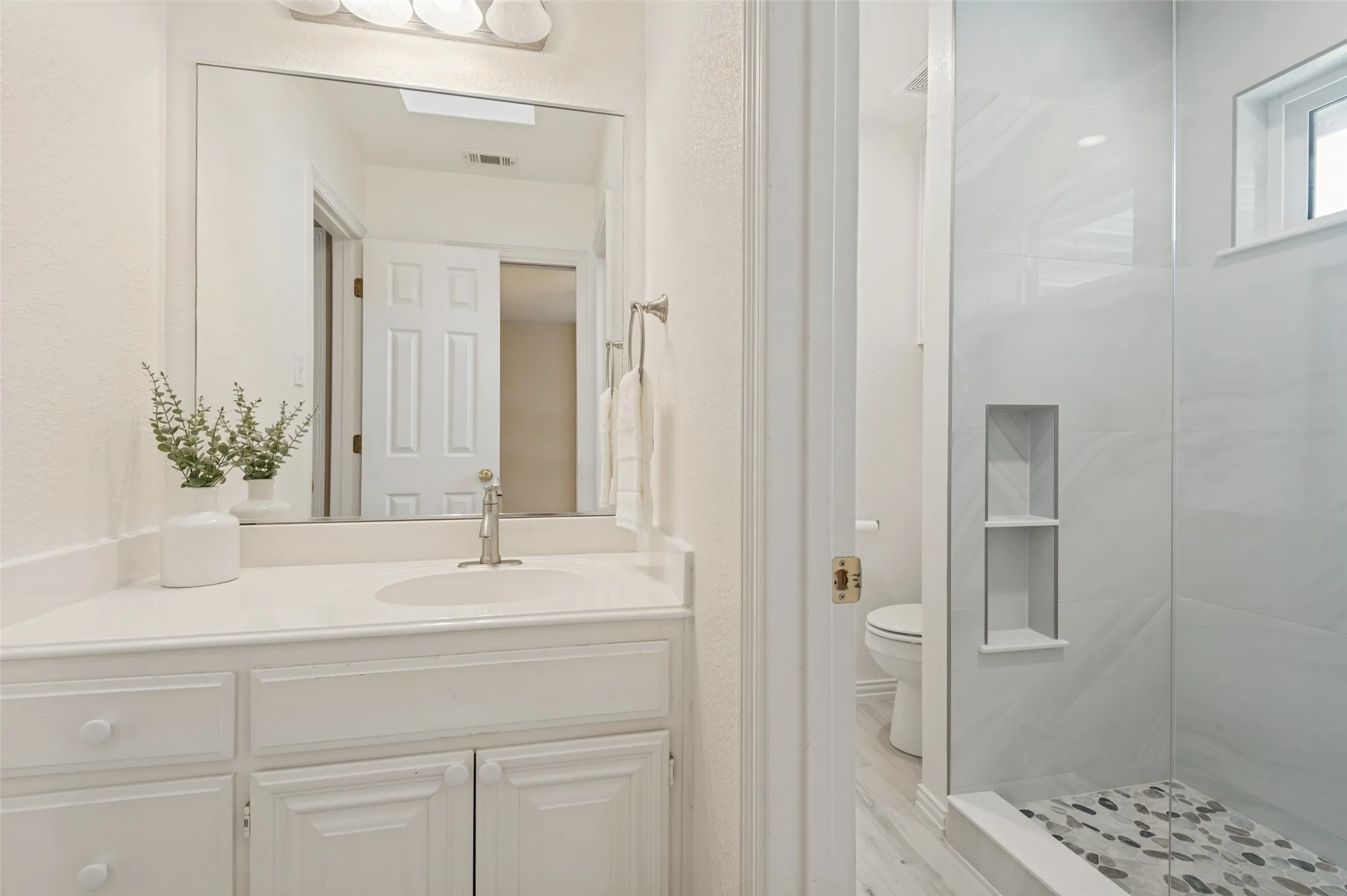 Bathroom vanity with a white countertop and oval under-mount sink, large wall-mounted mirror, walk-in shower with tile surround, built-in shower niche, and stone-tiled shower floor