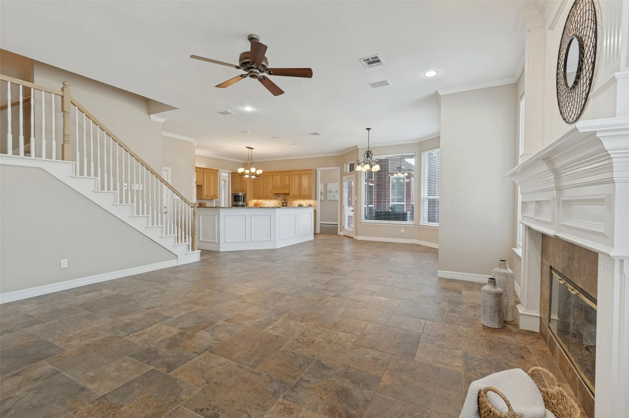 Open-concept interior featuring large-format stone tile flooring, a fireplace with a white millwork mantel, wooden staircase with white balusters, and a kitchen island with white cabinetry
