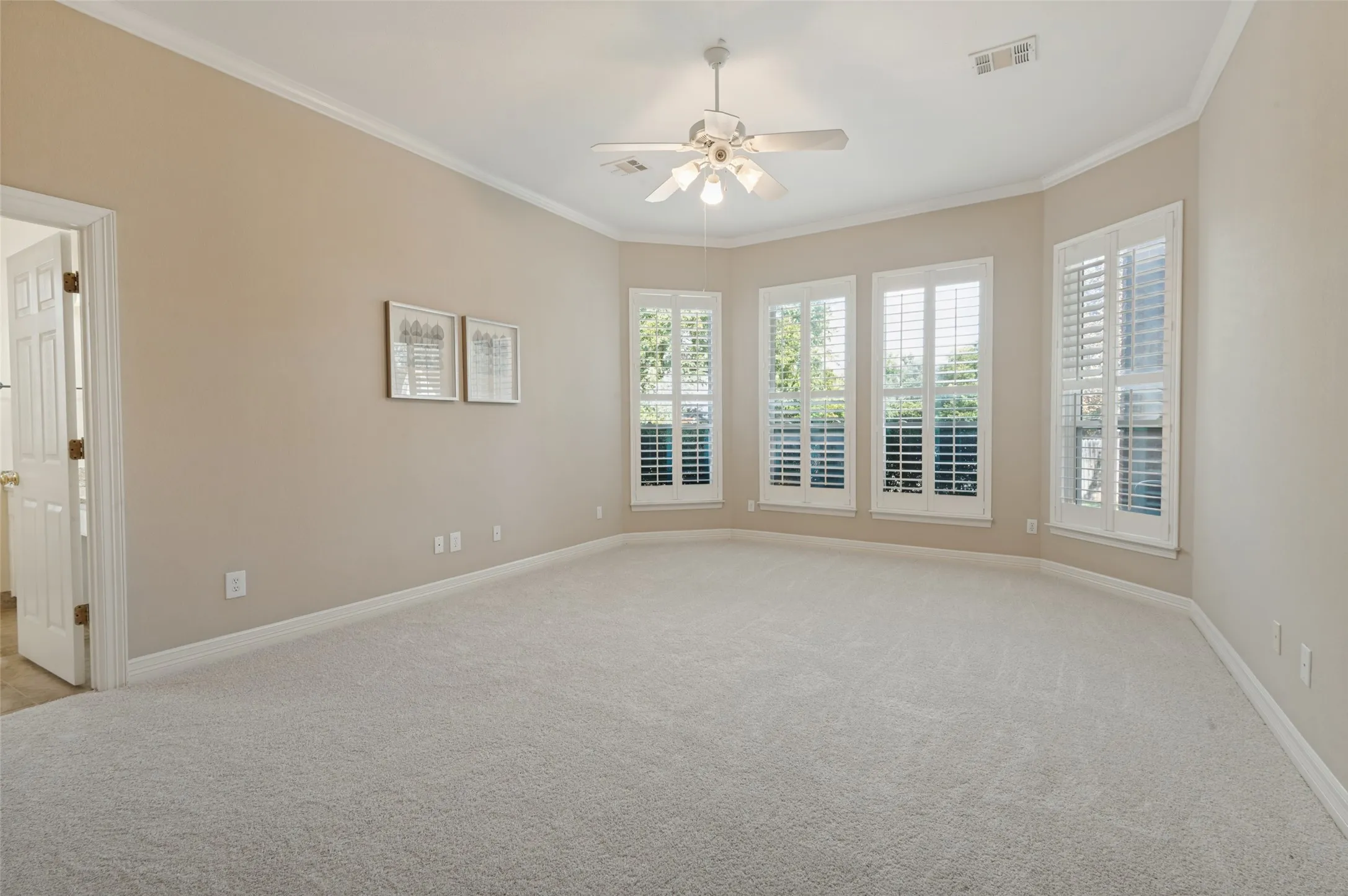Spacious room featuring neutral carpeting, a central ceiling fan with integrated lighting, and bay-style windows dressed with white plantation shutters