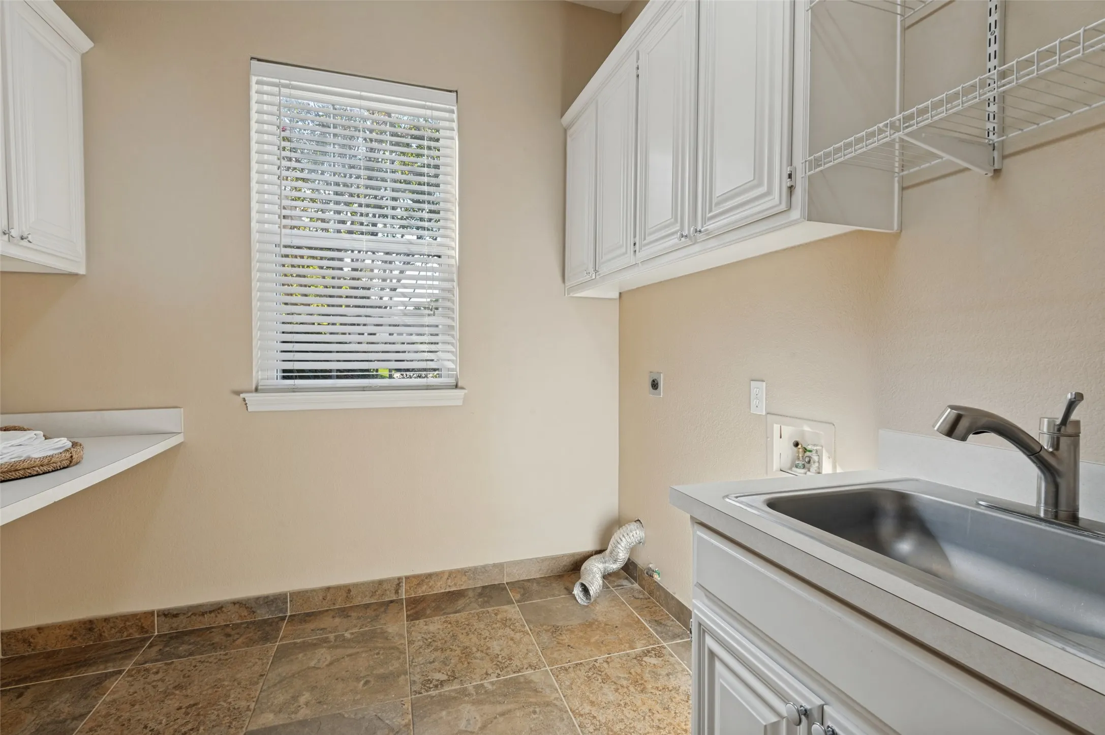 Laundry space featuring white shaker-style cabinetry, a stainless steel utility sink with a chrome faucet, stone-finish tile flooring, and a fixed laminate countertop
