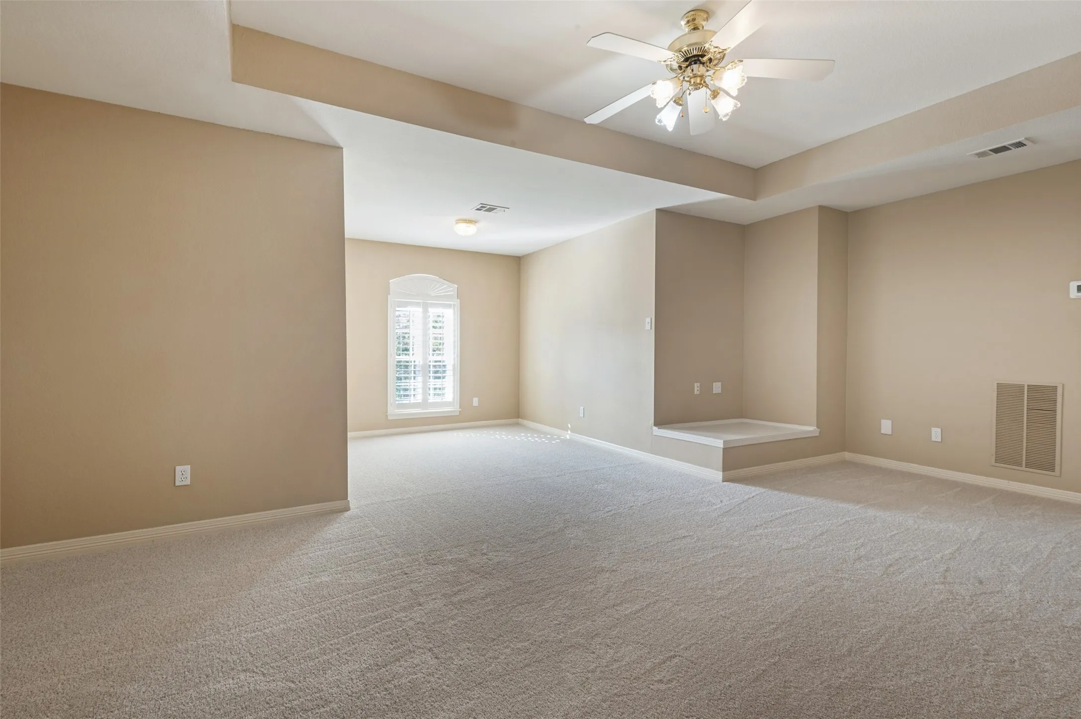 Spacious room featuring a tray ceiling, neutral wall-to-wall carpeting, an arched window with plantation shutters, a built-in architectural alcove, and a multi-blade ceiling fan with integrated lighting