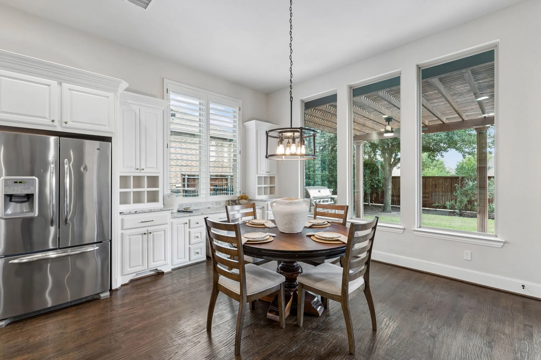 Eat-in kitchen featuring white cabinetry, wood-finish flooring, plantation shutters, and a suspended metal-frame light fixture with oversized windows overlooking a covered outdoor patio