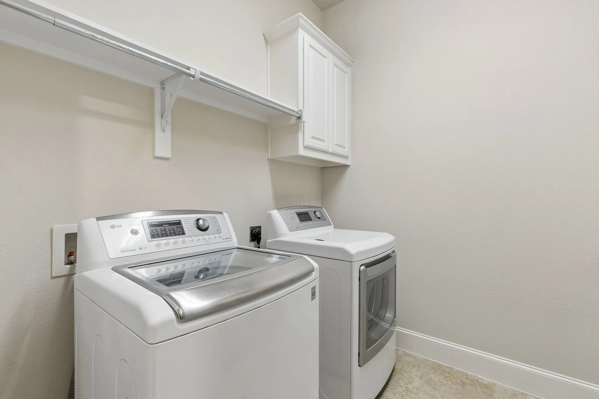 Utility space featuring white shaker-style upper cabinetry, a sturdy wall-mounted shelf with integrated clothing rod, and neutral tile flooring