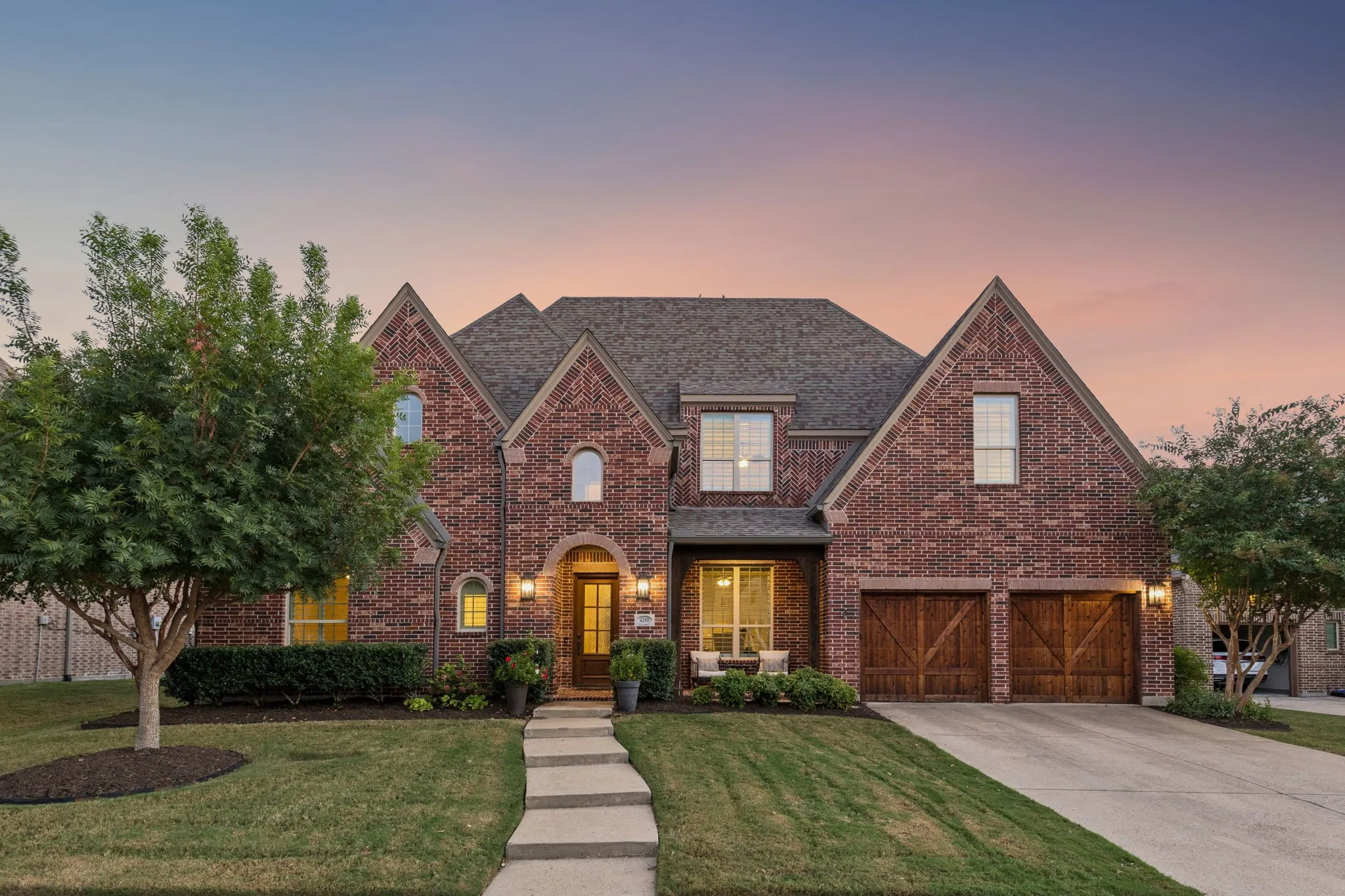 Multi-gabled brick exterior featuring wood-finished carriage-style garage doors, a stone walkway, and shingled roofing