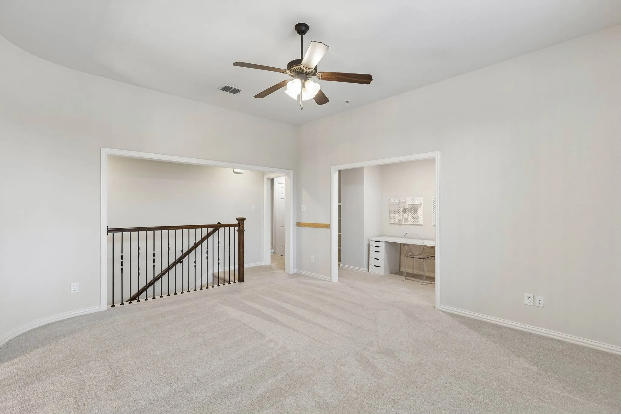 Neutral carpeted interior featuring high ceilings, a ceiling fan with integrated light fixture, metal baluster railing, and built-in workspace