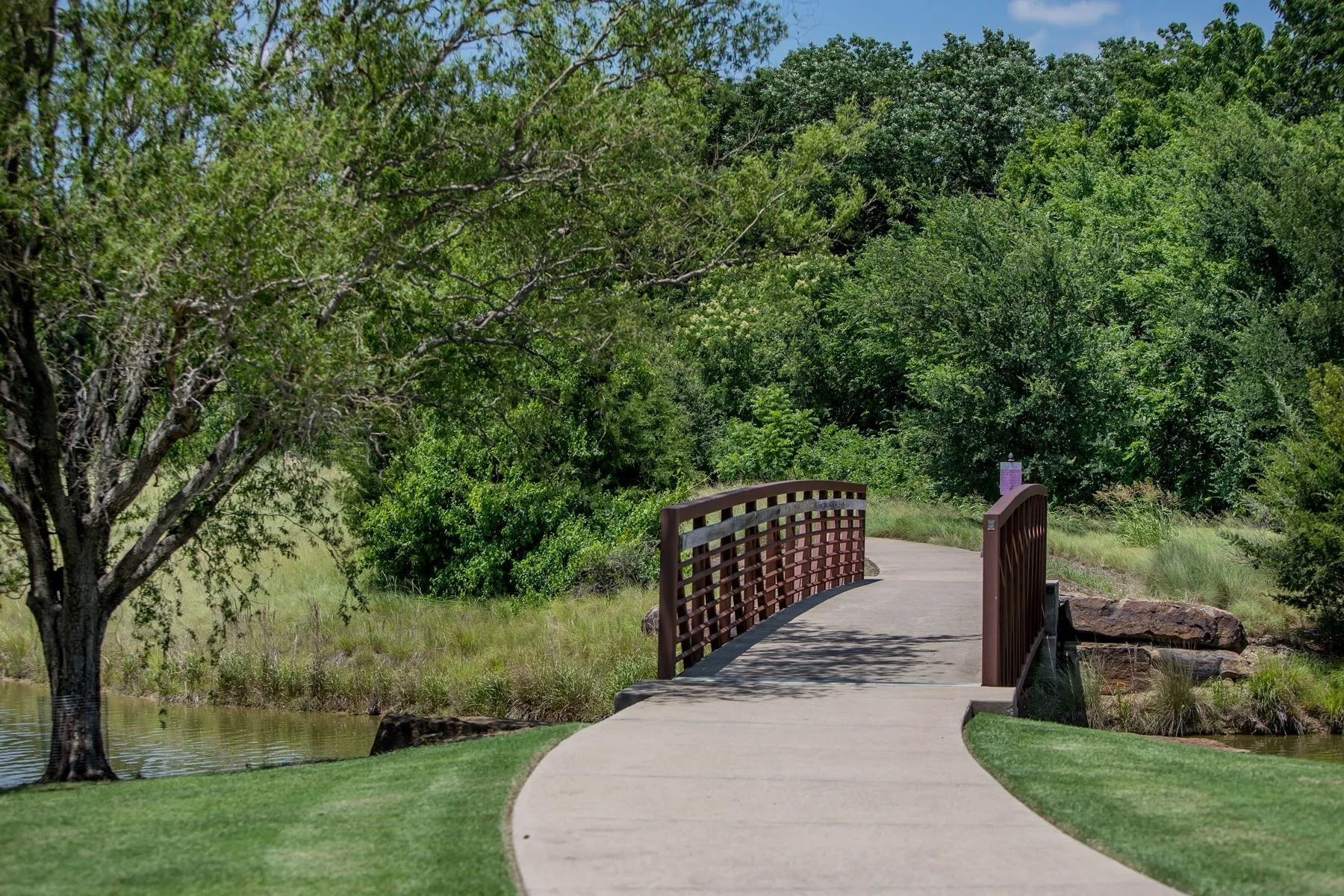 Concrete pathway featuring a metal bridge, stone retaining walls, manicured lawn, and mature trees