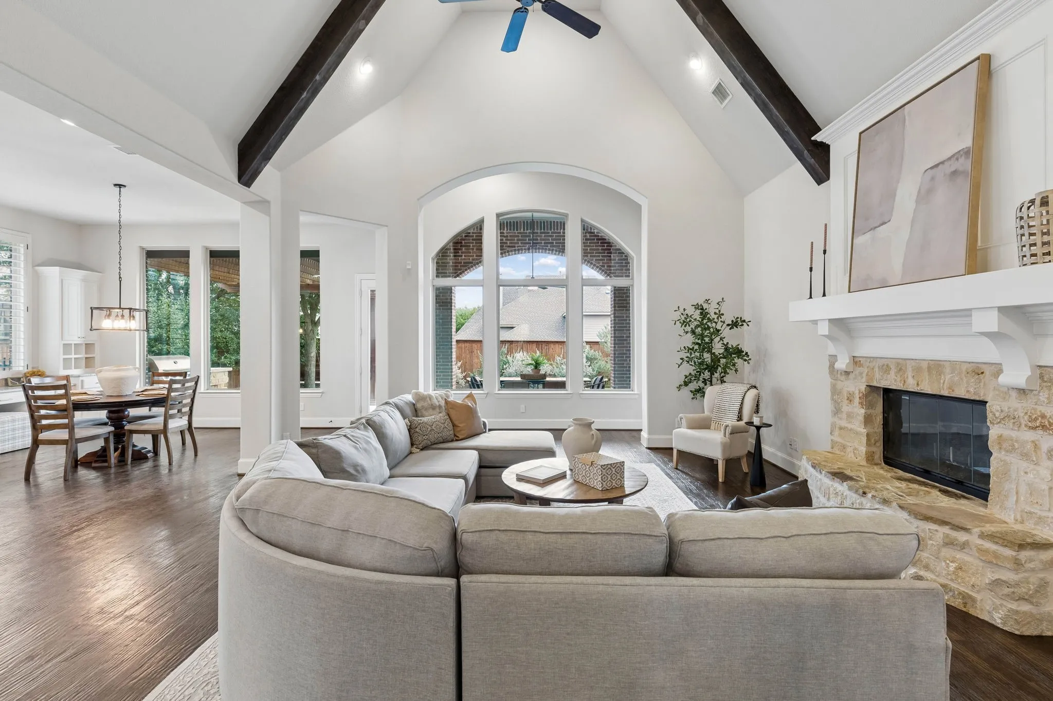 Vaulted ceiling with exposed decorative beams, stone-surround fireplace with a wooden mantel, arched floor-to-ceiling windows, and wood-finish flooring