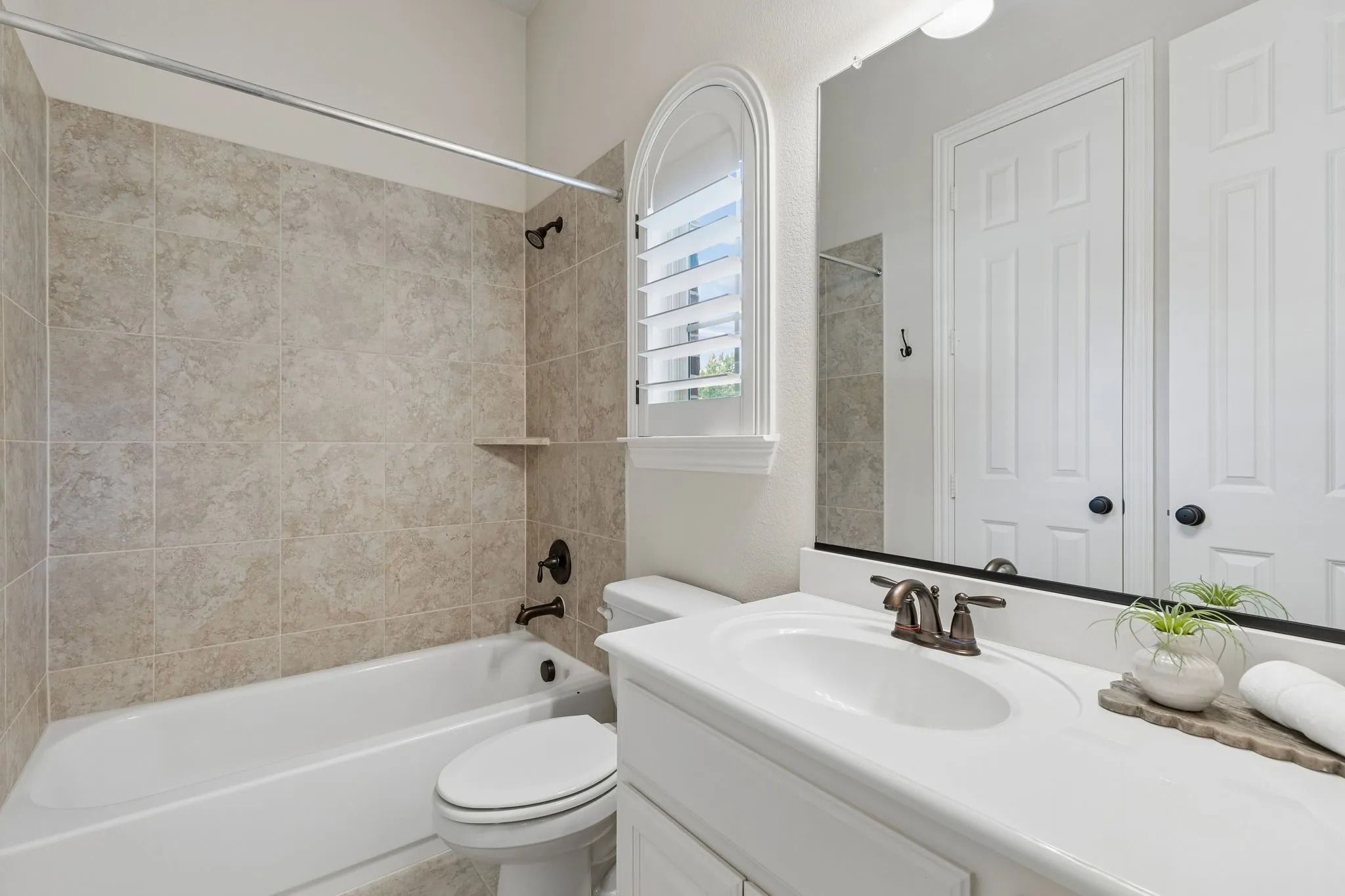Bathroom featuring a vanity with an integrated countertop, arched window with plantation shutters, tub-shower combination with tile surround, and oil-rubbed bronze fixtures