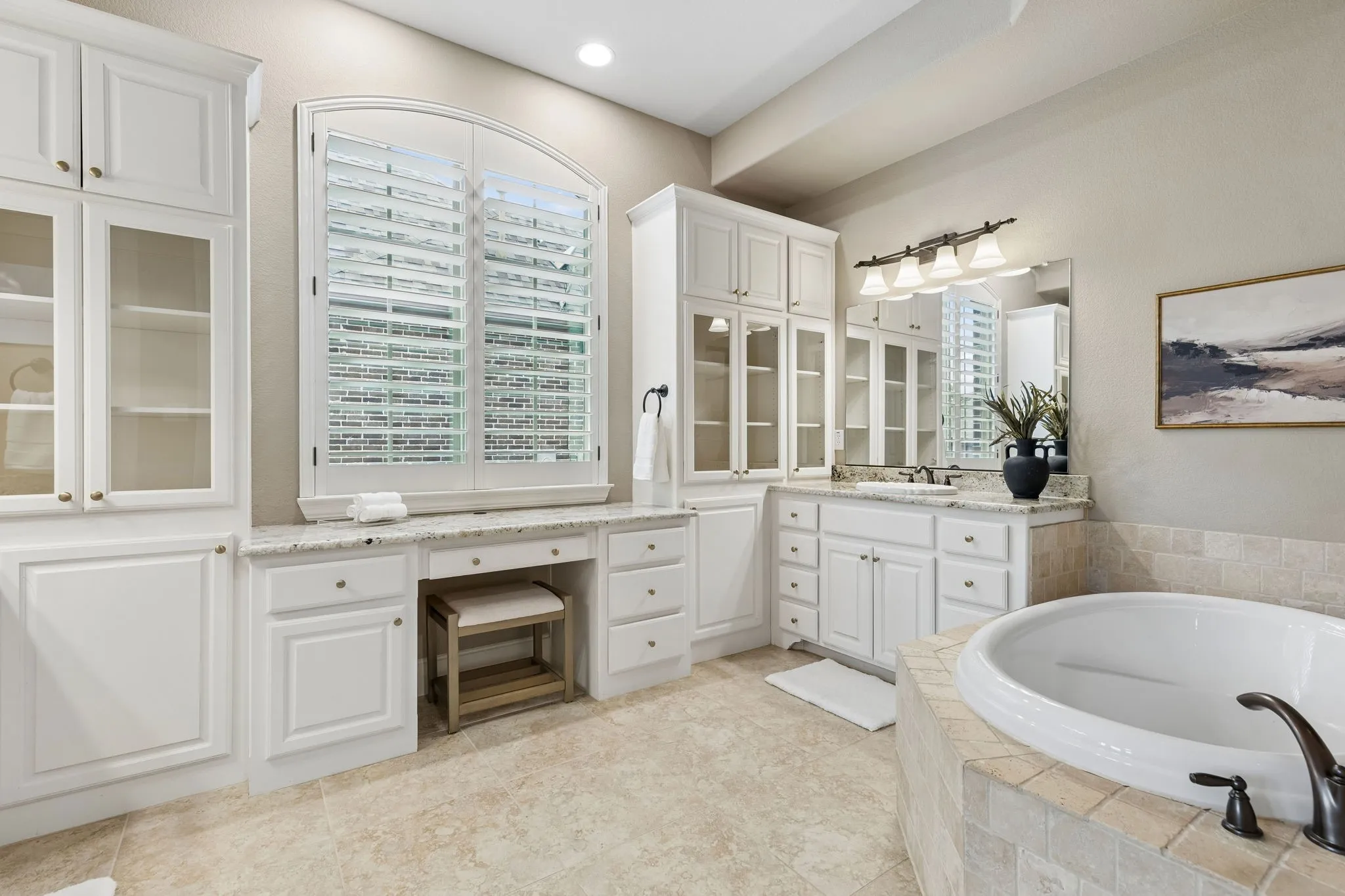 White cabinetry with glass-front uppers, stone-surfaced vanity and dressing area, arched window with plantation shutters, tile-surrounded soaking tub, and recessed lighting