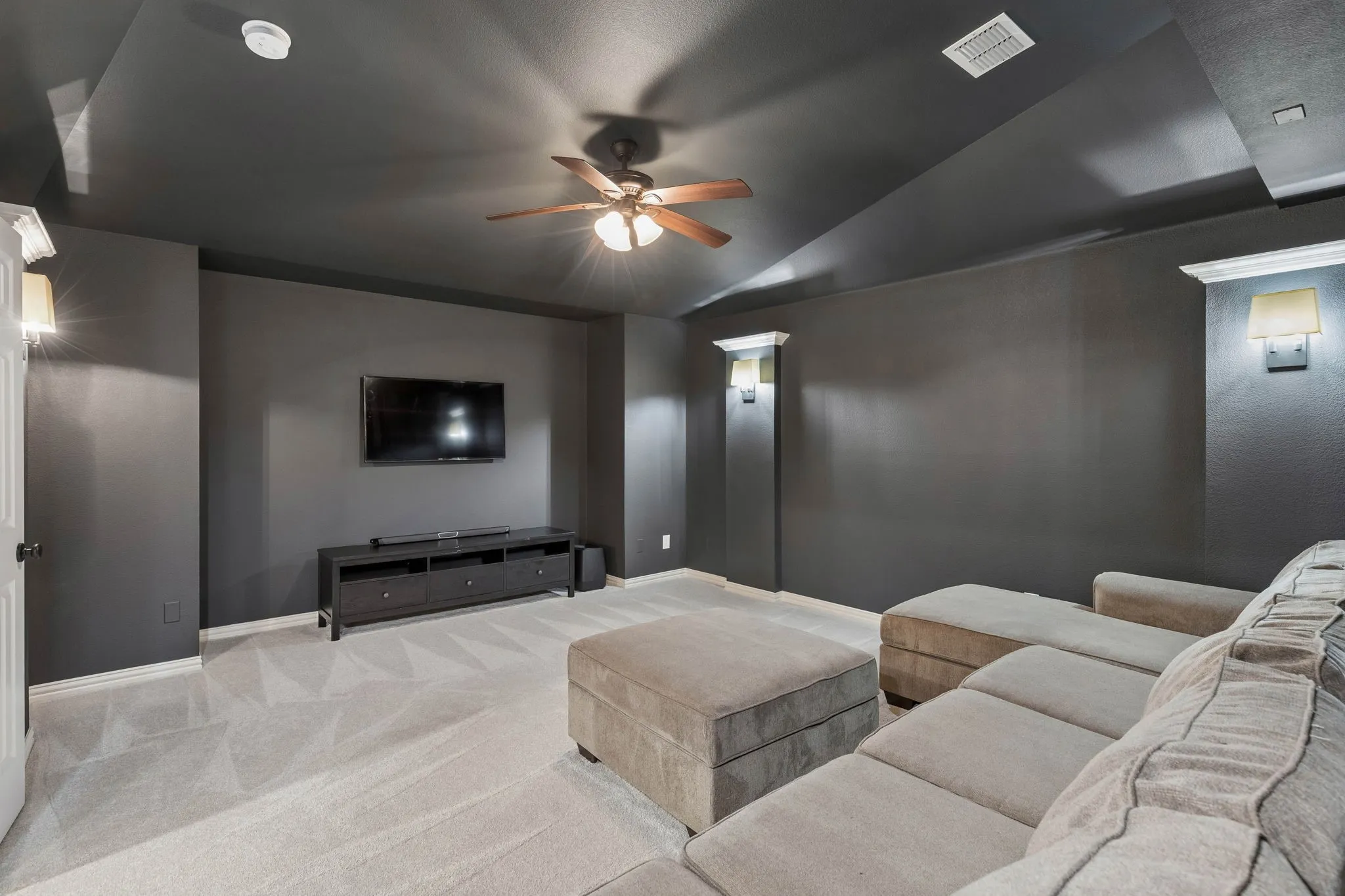 Tray ceiling with a central multi-blade ceiling fan, wall-mounted sconce lighting, and neutral carpeting