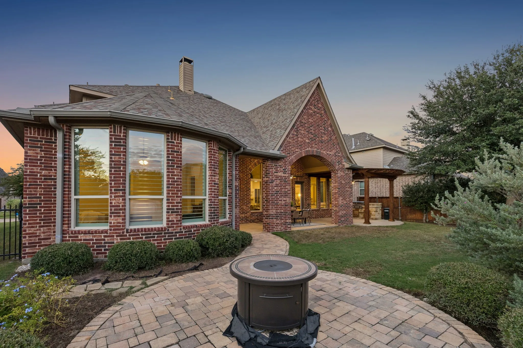 Brick exterior featuring a multi-faceted roofline, stone-paver circular patio with a built-in fire feature, covered porch, and a wood-beam pergola