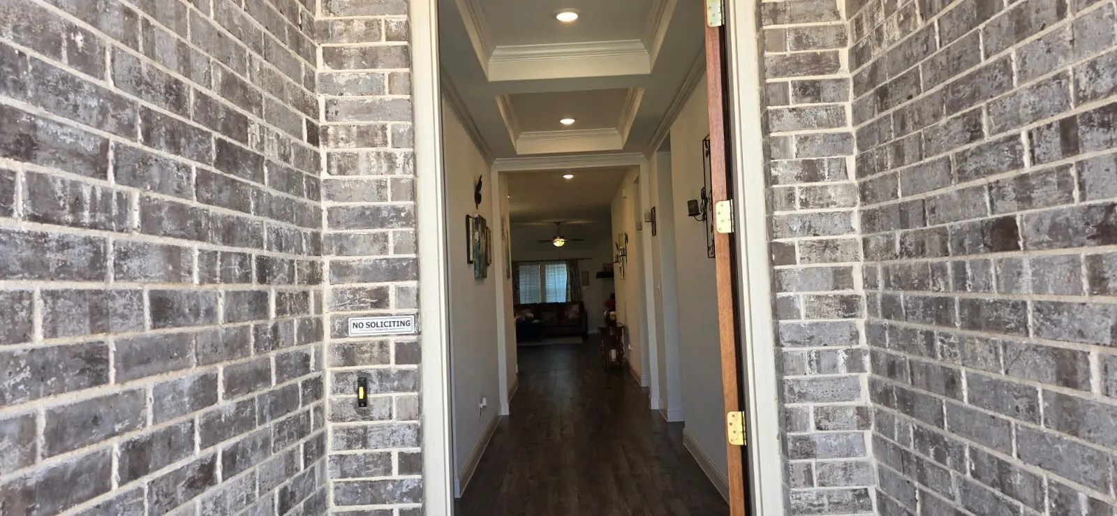 Gray brick exterior with a wood-finish entry door, leading to a hallway with tray ceilings and recessed lighting