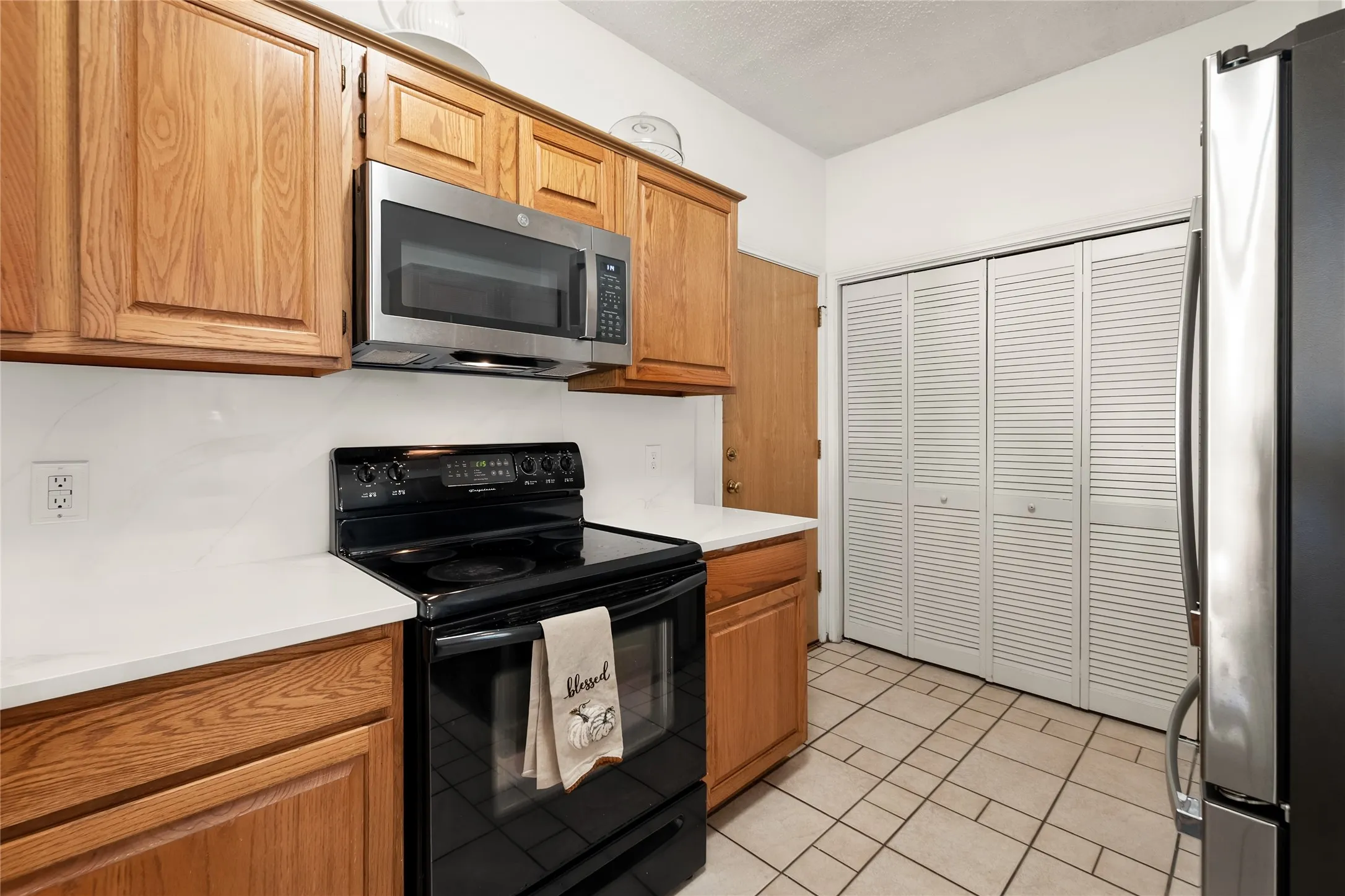 Kitchen featuring medium-tone wood cabinetry, solid-surface countertops, built-in stainless steel microwave, black electric range, and tiled flooring