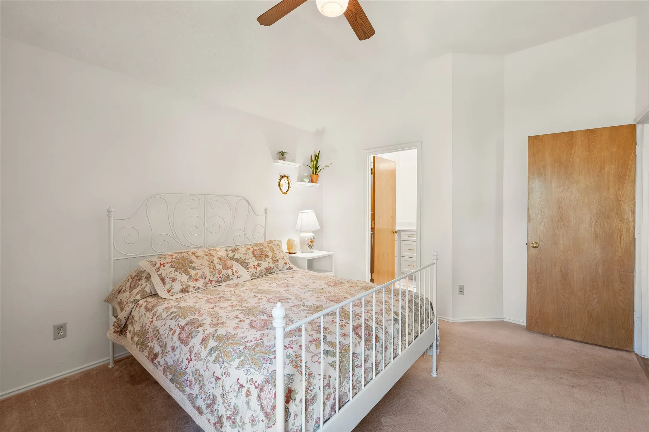 High-ceiling room featuring neutral carpeting, a central ceiling fan with an integrated light fixture, two built-in wall-mounted shelves, and a solid wood-finish interior door
