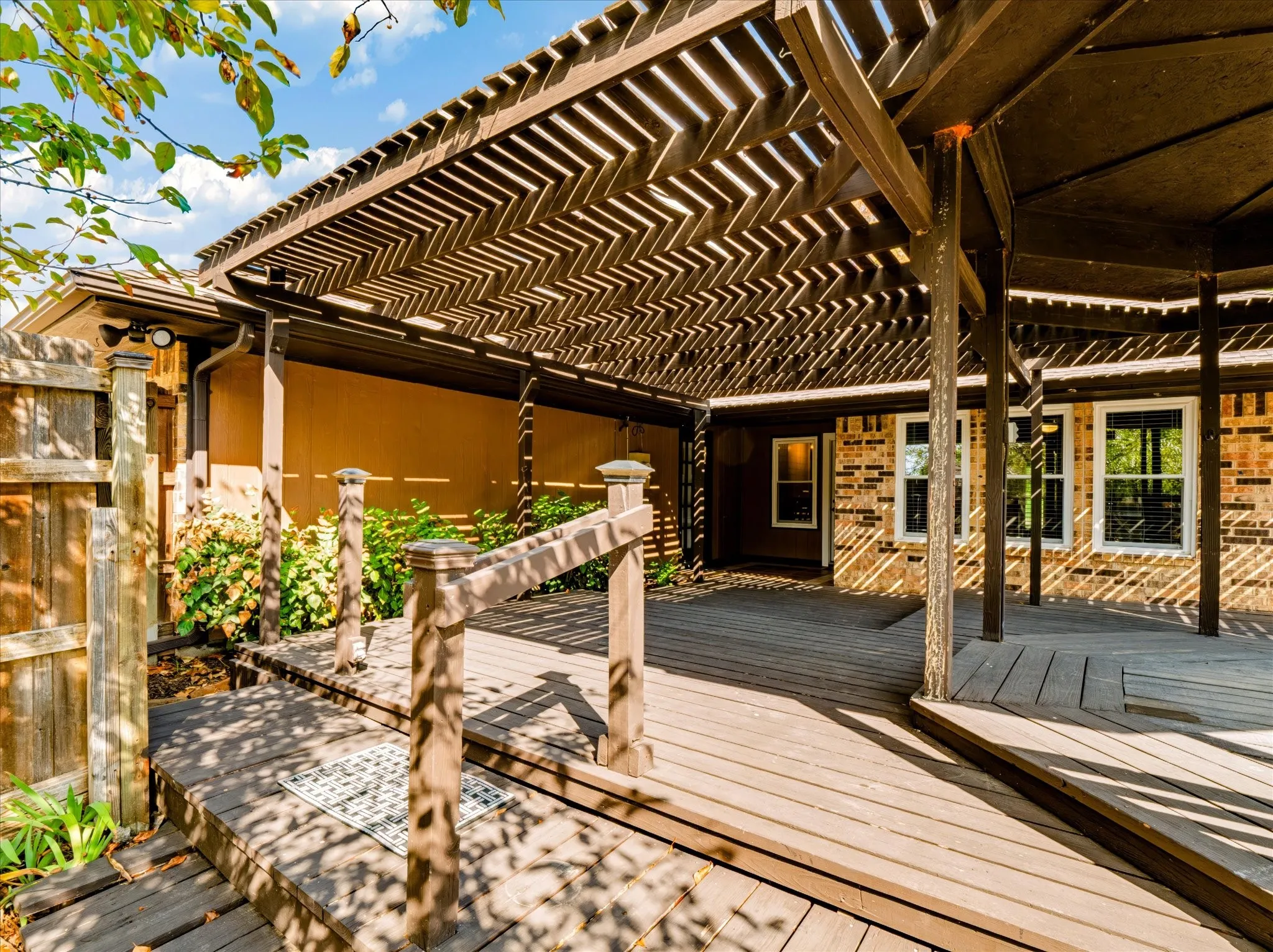 Multi-level wood deck featuring a slatted timber pergola, recessed architectural lighting, brick exterior siding, and a wooden handrail
