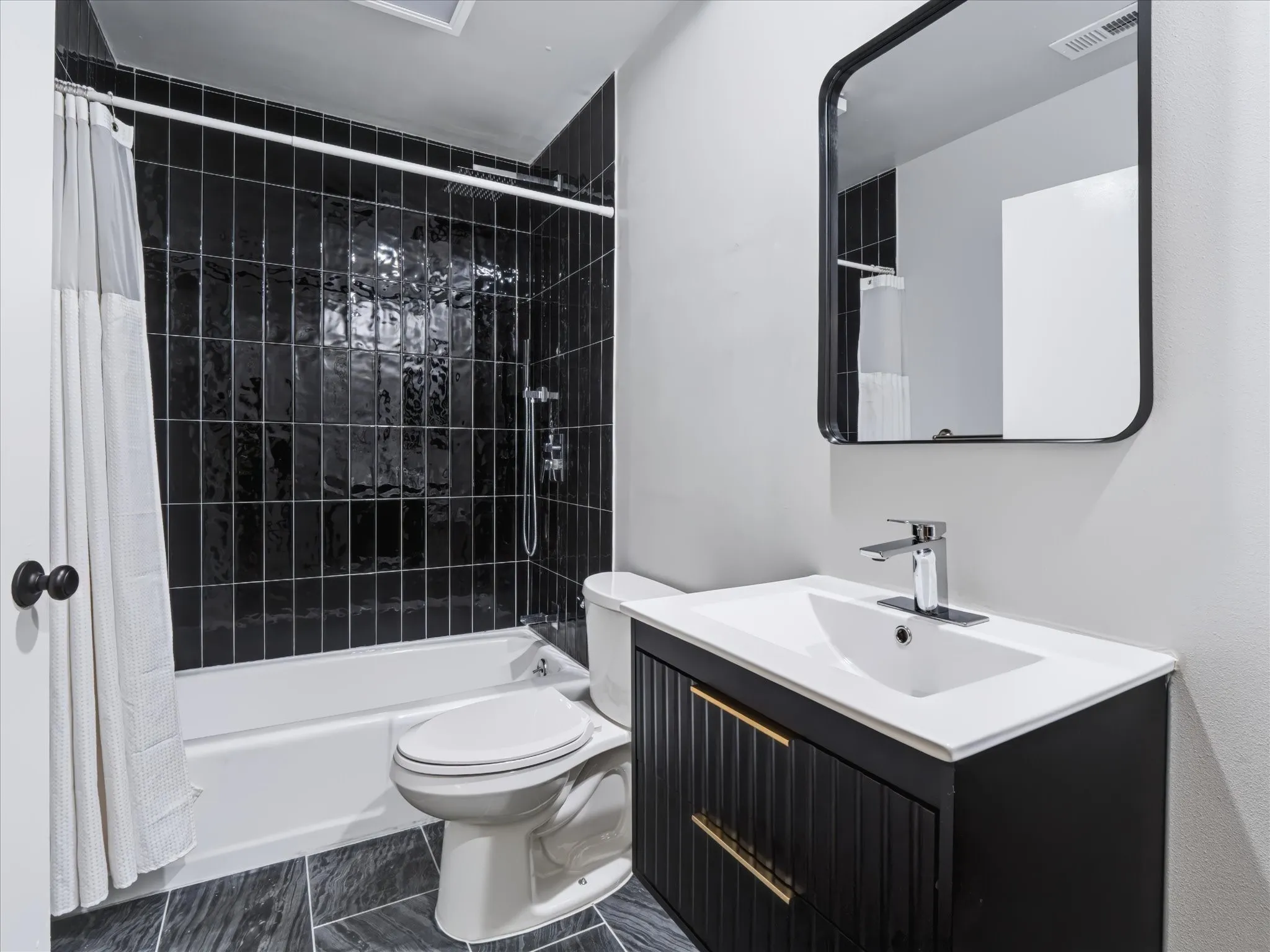 Bathroom featuring vertical black wall tiles in the shower area, dark-finished vanity with gold-tone hardware, white countertop with integrated basin, polished metal faucet, and black-framed wall mirror