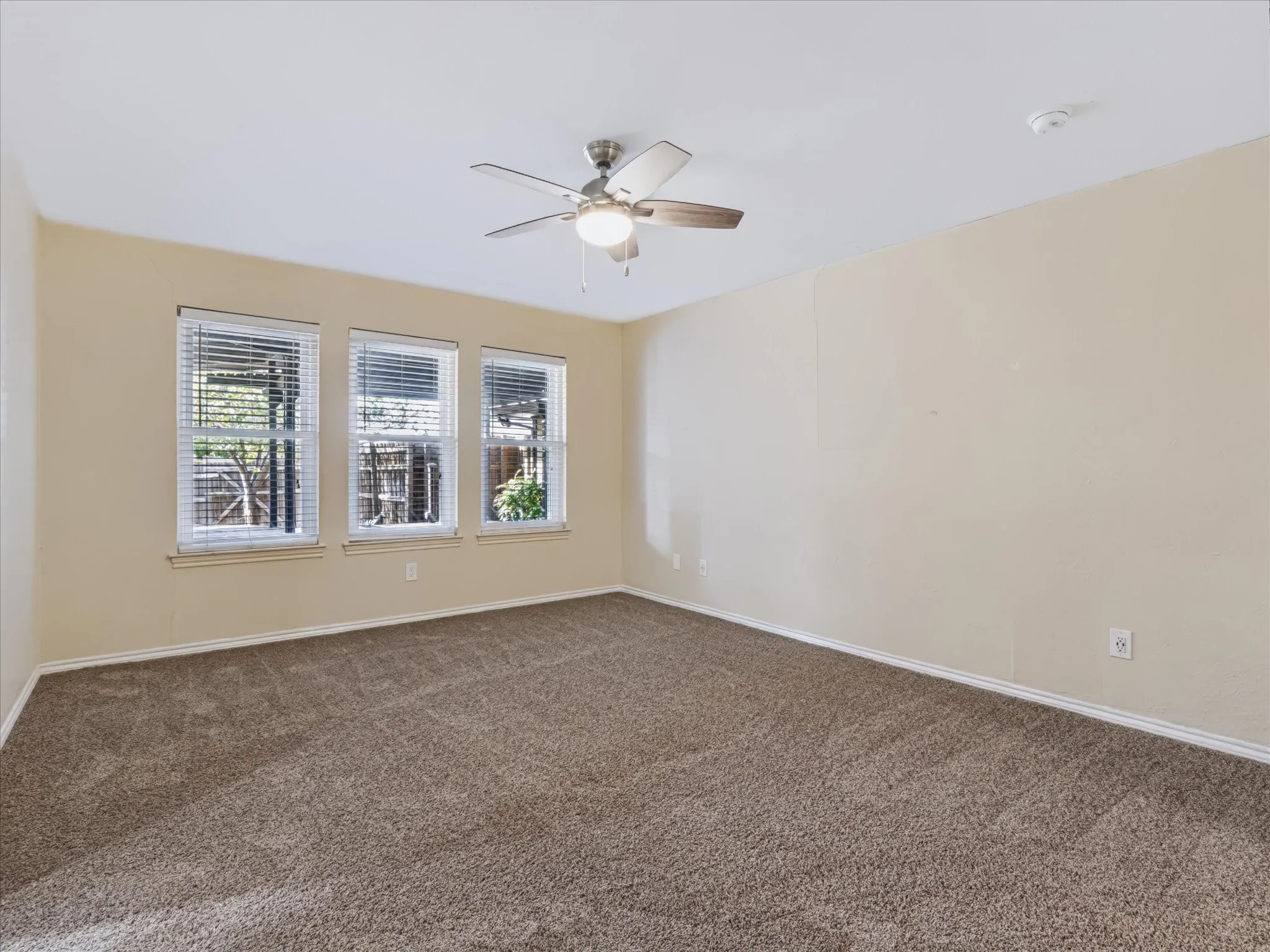 Neutral-toned interior space featuring wall-to-wall carpeting, three multi-pane windows with horizontal blinds, and a central ceiling fan with an integrated light fixture