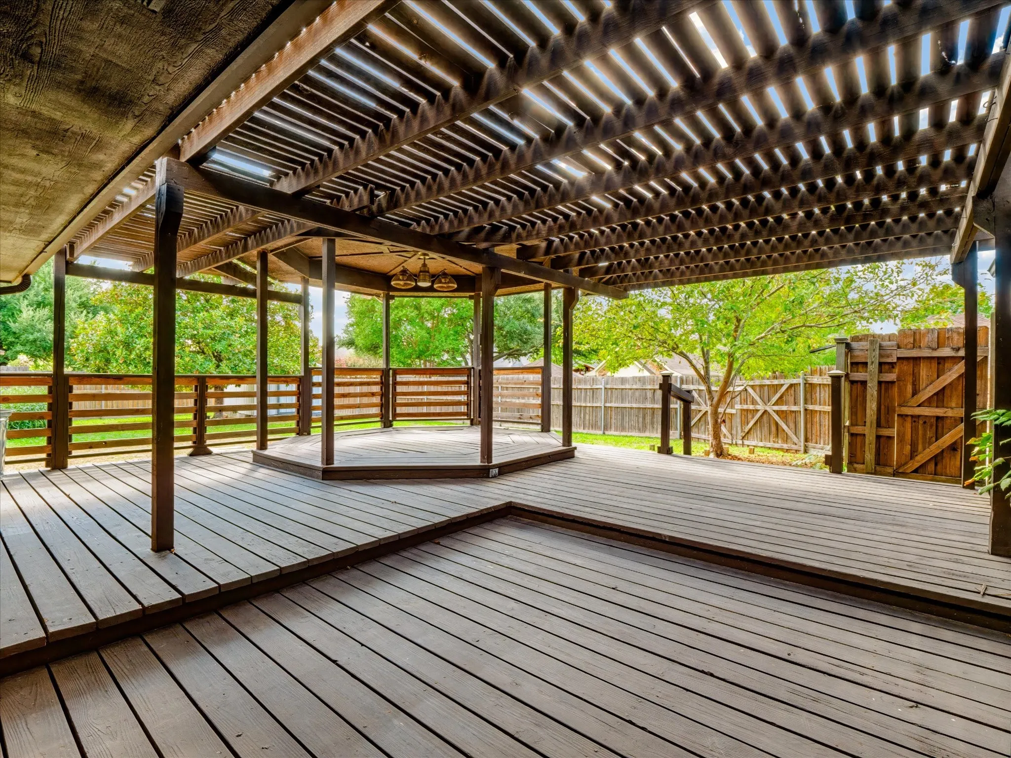 Multi-level wooden deck featuring a tiered hexagonal pavilion, overhead pergola, wooden perimeter railings, and a fenced backyard boundary